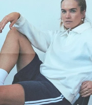 Woman sitting with legs crossed wearing a white hoodie and dark shorts. Los Angeles premiere hair and makeup artist, Amber Rose.