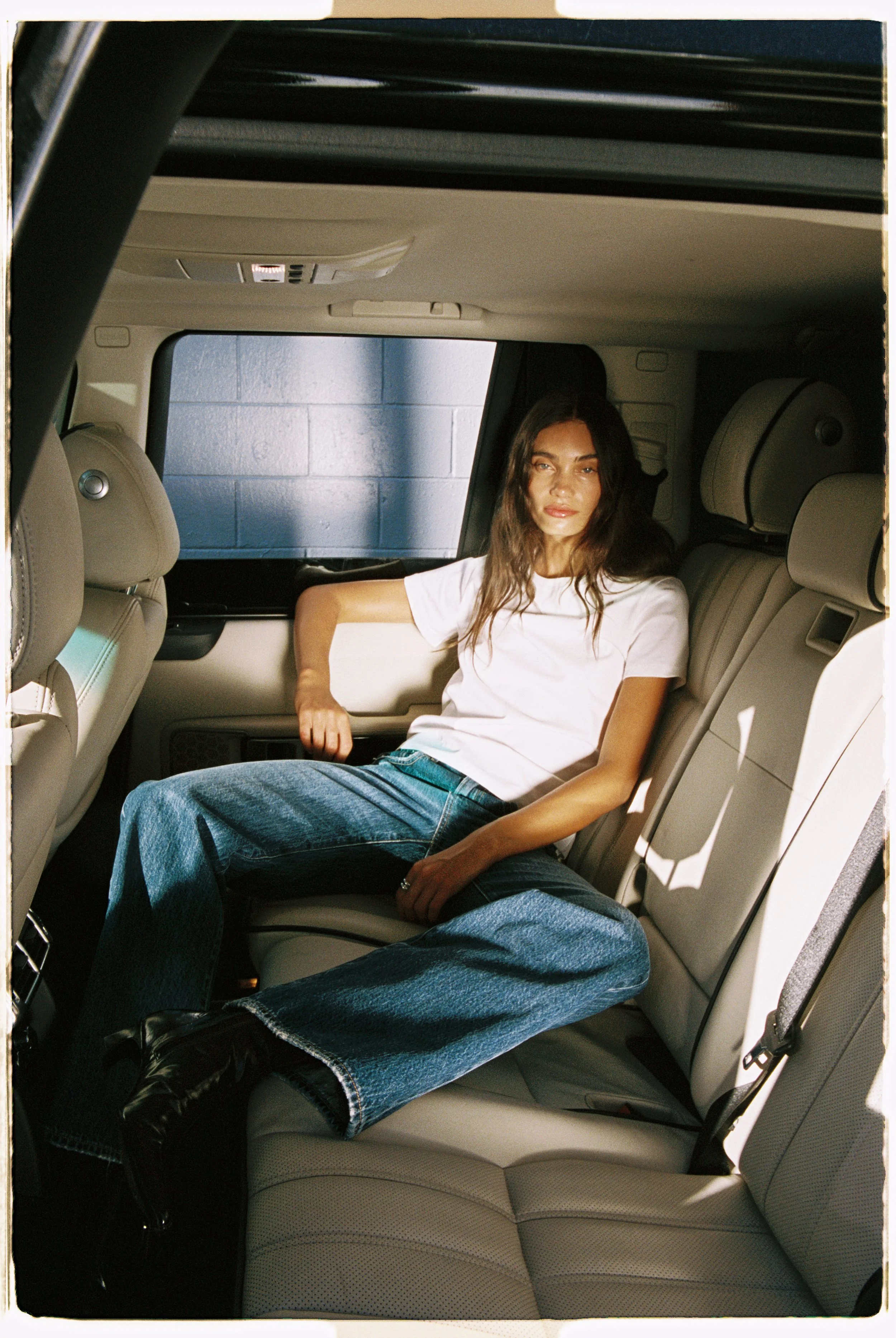 Young woman with long dark hair and light skin wearing a white t-shirt and blue jeans, sitting in the back seat of a car with beige interior, looking at the camera.