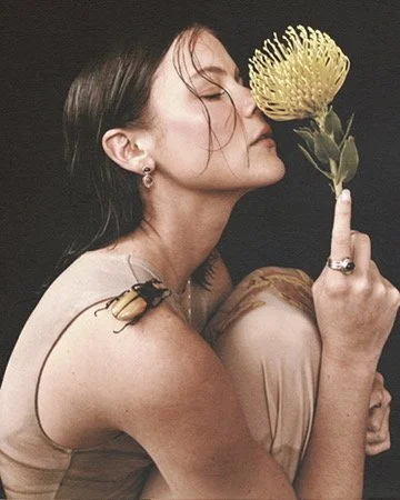 Nature-inspired editorial shot of a model smelling a yellow protea flower with a beetle brooch on her shoulder, hair and makeup by Los Angeles artist Amber Rose.
