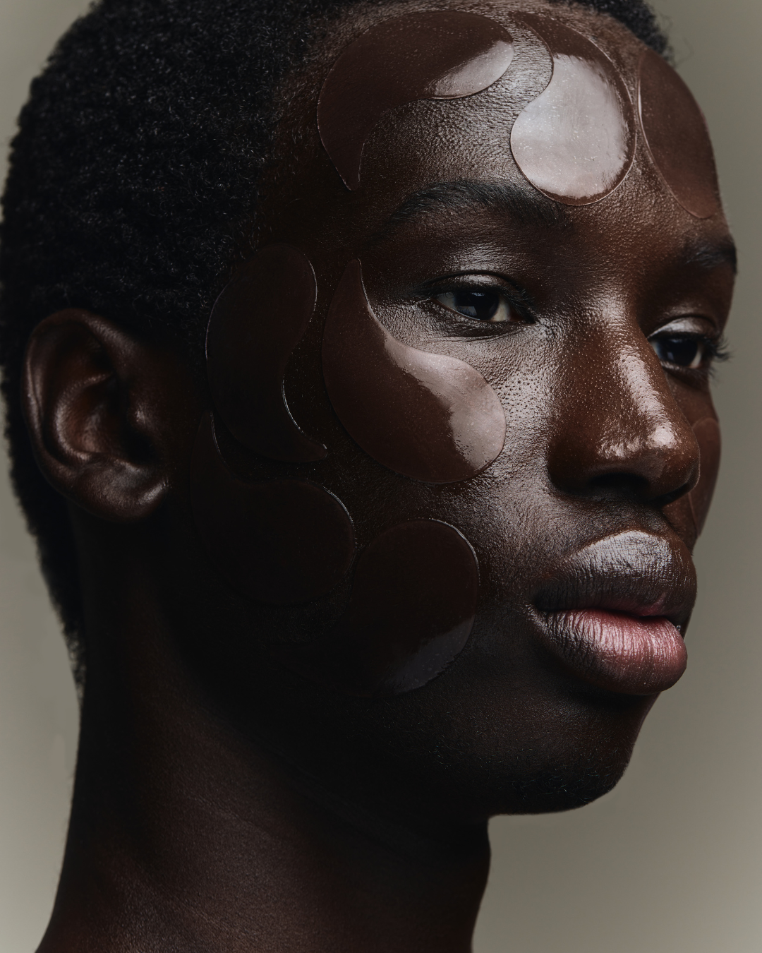 Close-up of a model's face with dark skin, short curly hair, and decorative brown adhesive eye patches on their face. Beauty campaign.
