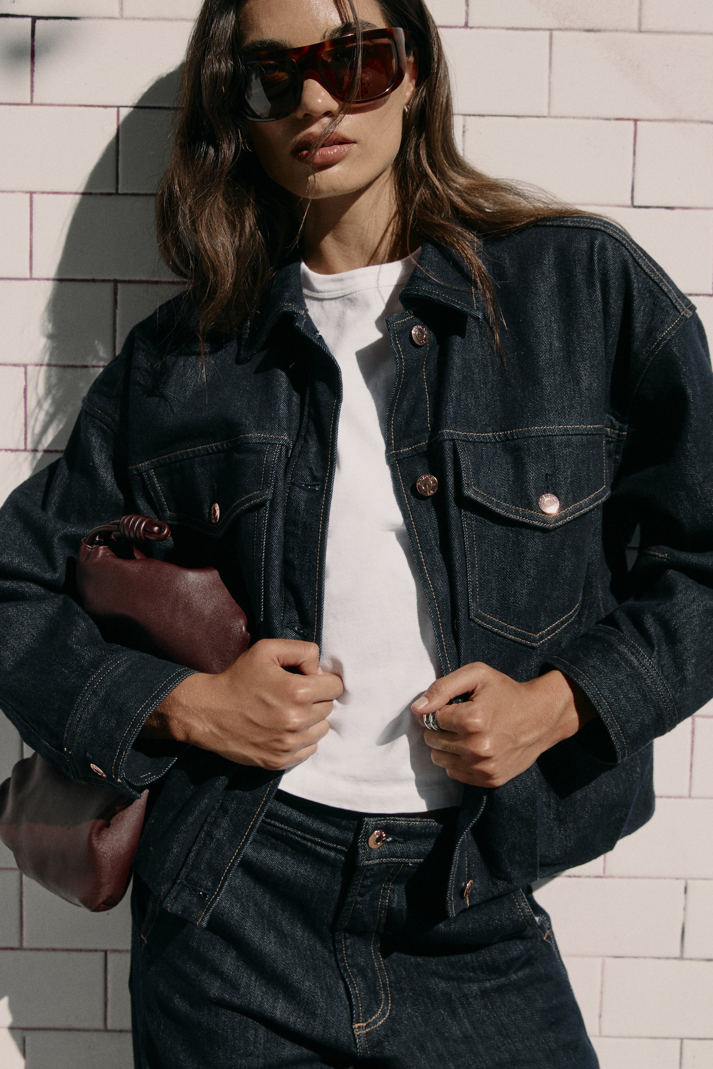 A woman wearing large sunglasses and a denim jacket, standing in front of a pink brick wall.