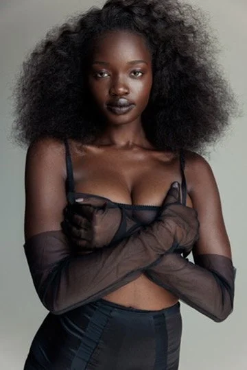 A woman with big curly hair wearing black gloves and a black lingerie top, looking at the camera against a neutral background. Hair and makeup by Amber Rose.