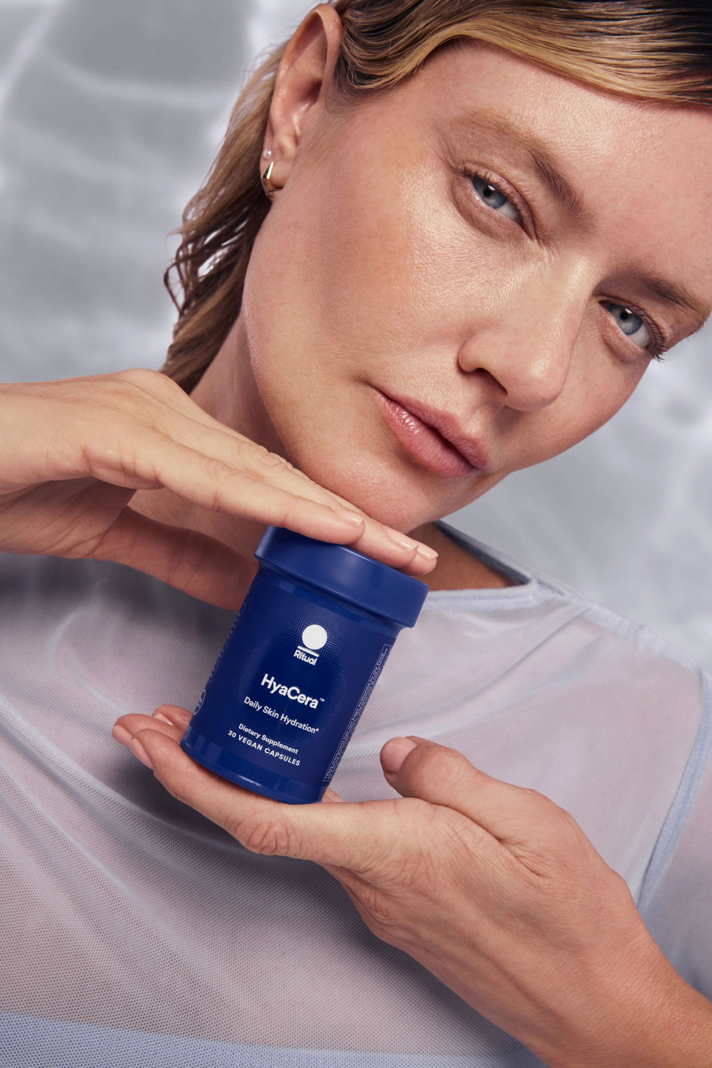 A woman with blonde hair and blue eyes holding a blue bottle of Hyacera Daily Skin Hydration dietary supplements close to her face and smiling gently. Hair and makeup by Amber Rose.