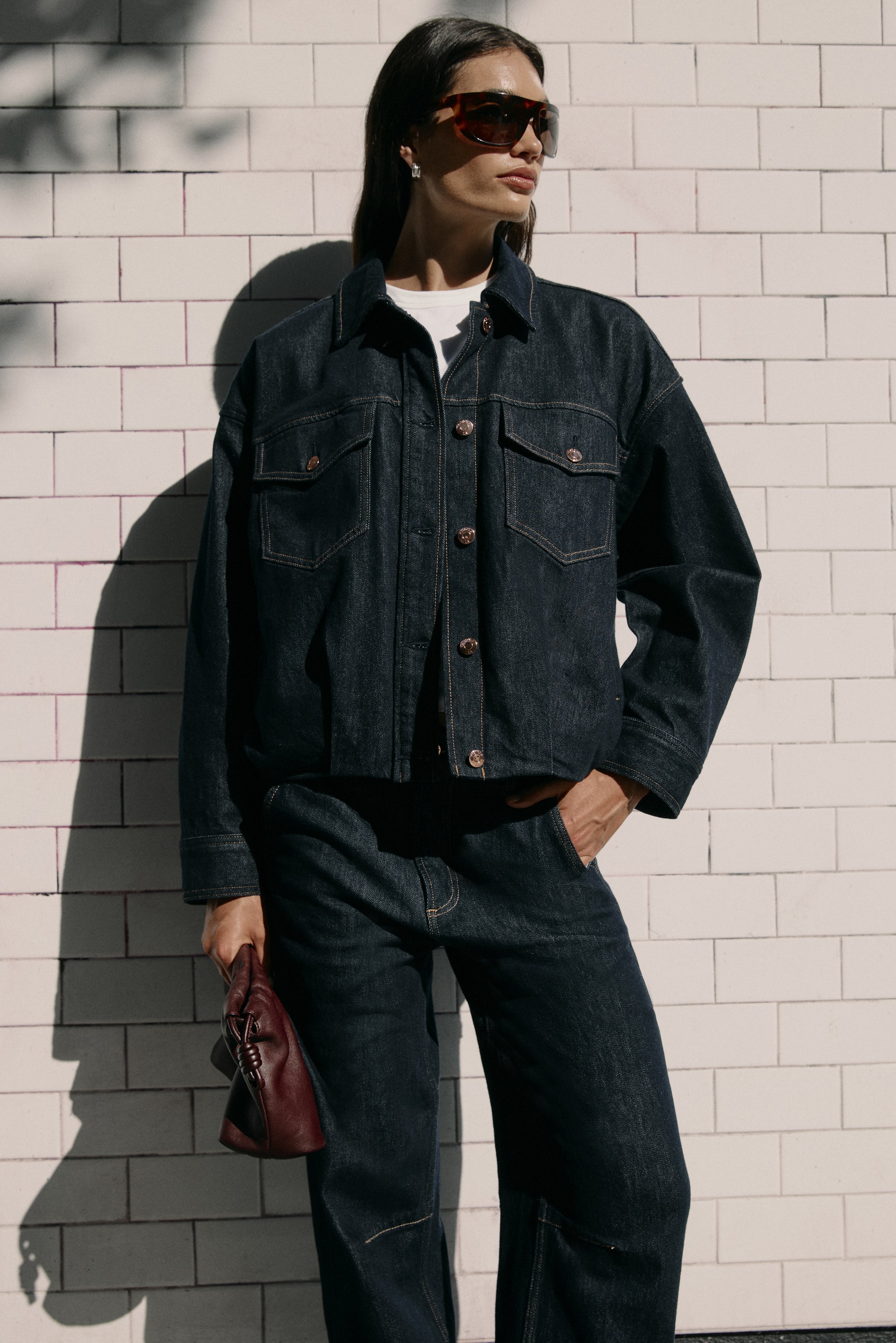 Woman wearing large sunglasses, dark denim jacket, and dark jeans, standing against a white brick wall, holding a burgundy handbag.