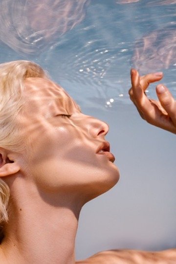 A woman with closed eyes and blond hair underwater, with a hand reaching towards her face. Hair and makeup by Amber Rose.