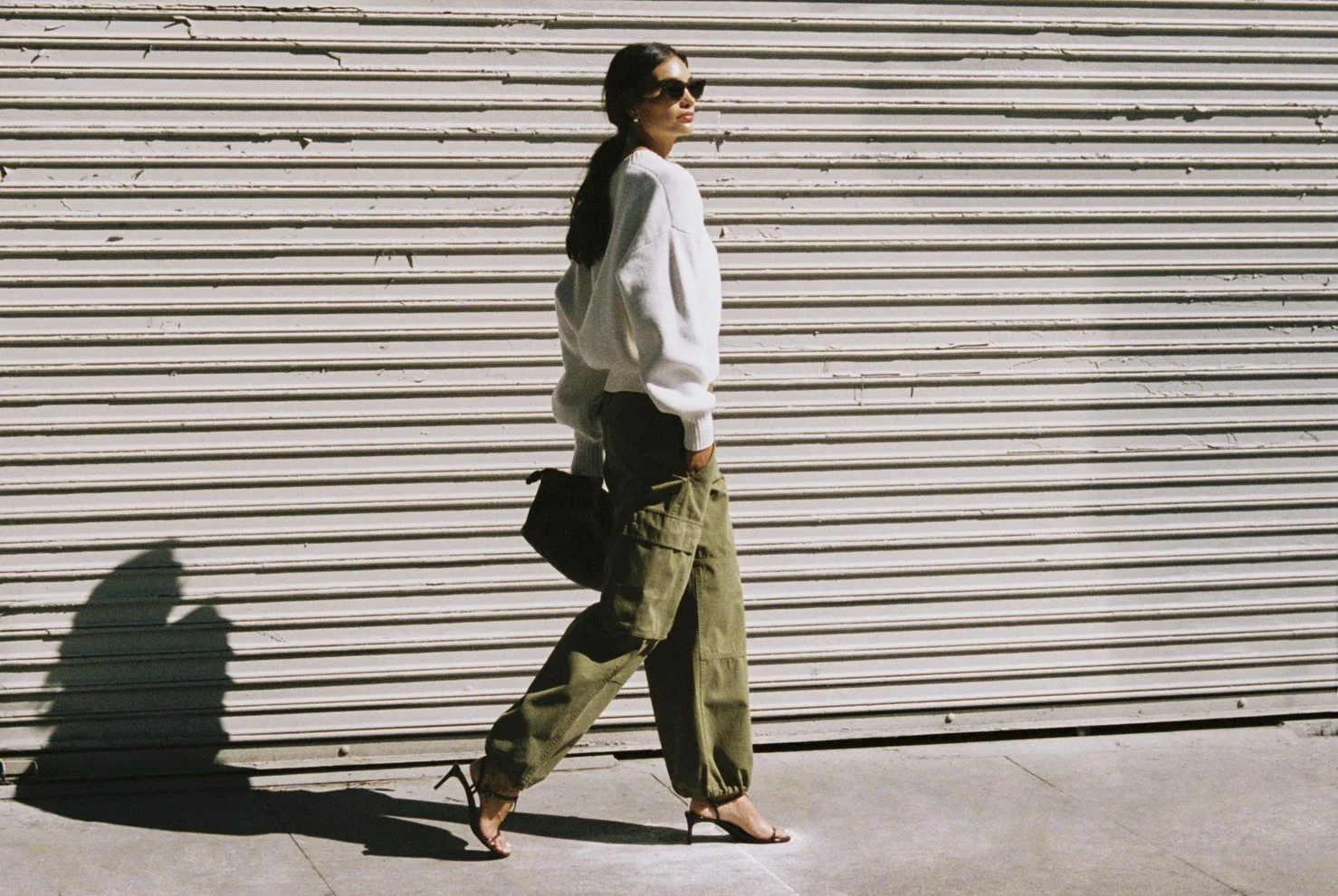A woman walking past a beige metal rolling shutter, wearing sunglasses, a white sweater, olive cargo pants, and high heels, with her shadow cast on the sidewalk. Hair and makeup by Amber Rose.
