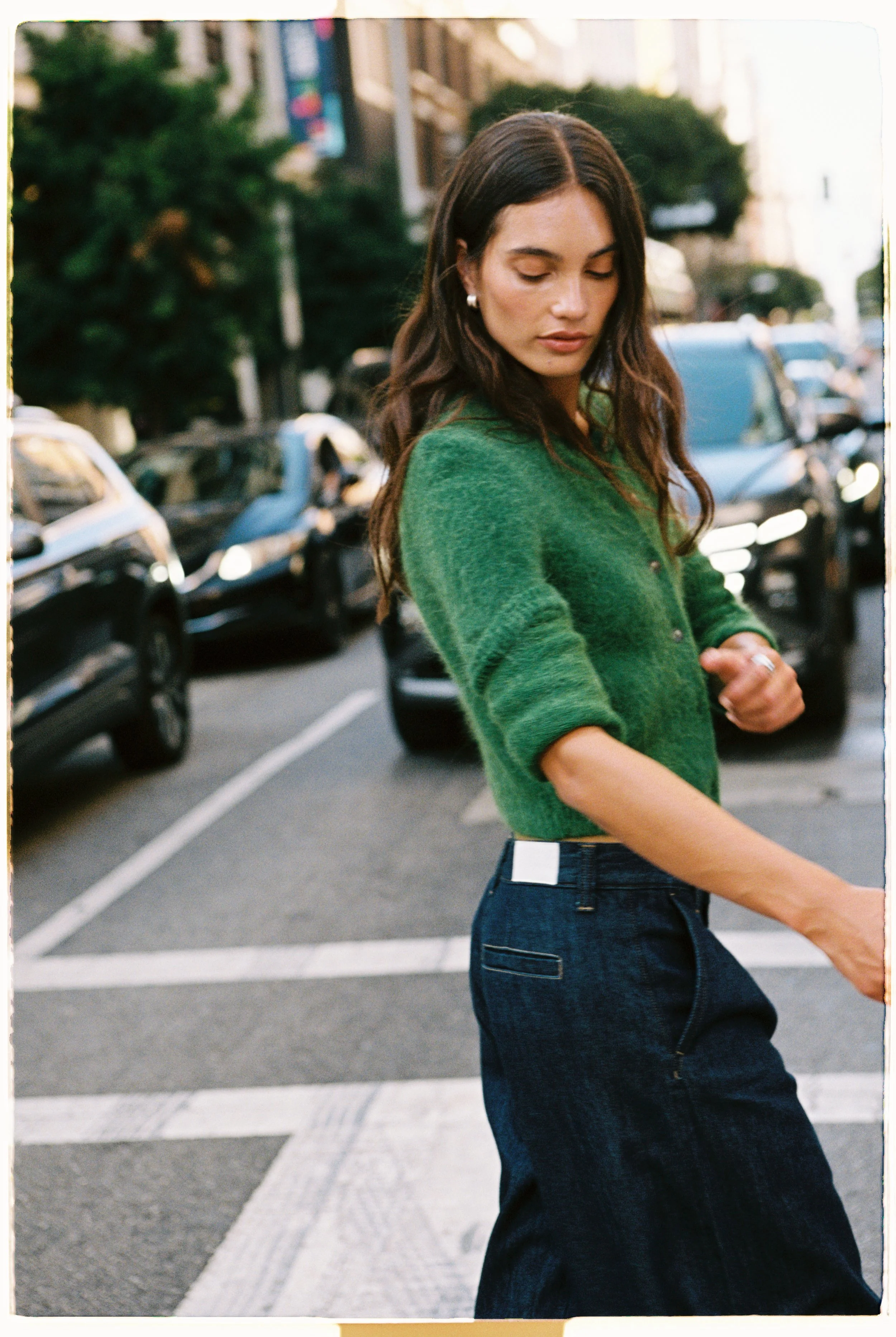 A woman wearing a green sweater and dark jeans walking in an urban street, with parked cars and buildings in the background.