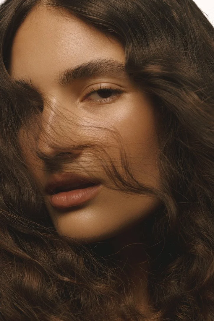 Close-up of a woman with long, wavy brown hair partially covering her face, one eye slightly squinted and lips slightly parted. Hair and makeup by Amber Rose.