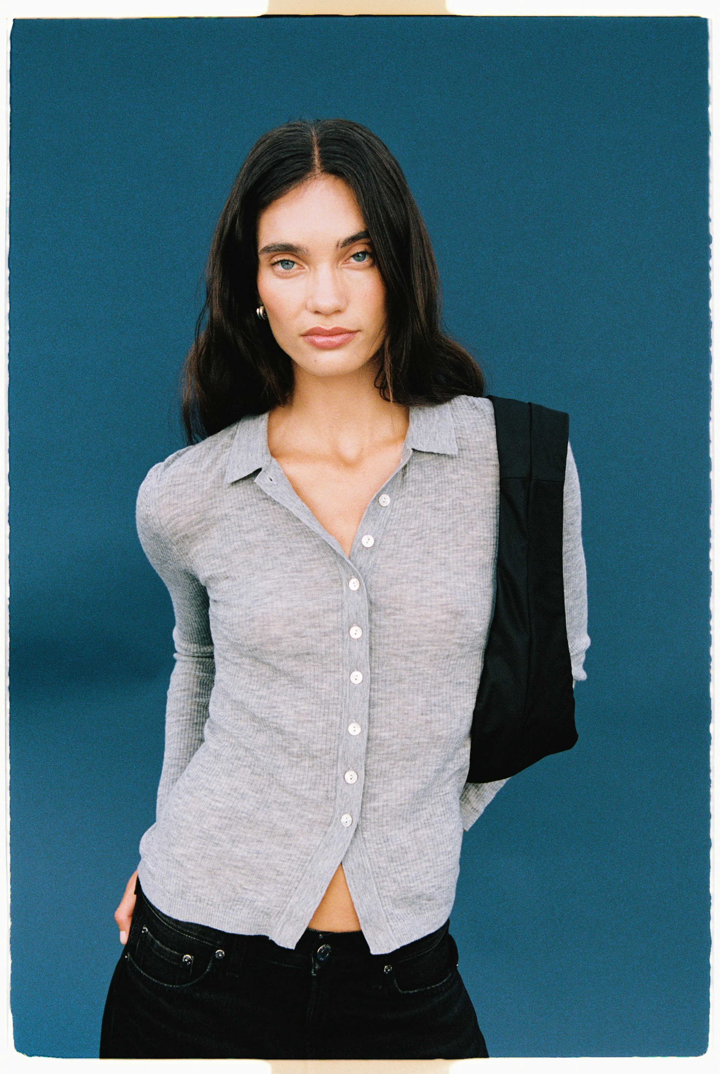 A young woman with dark hair and light blue eyes wearing a gray button-up shirt and black jeans, standing against a blue background.