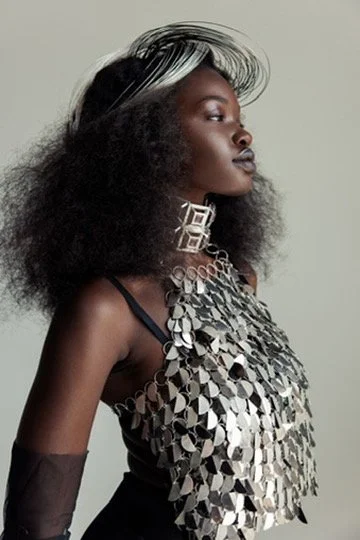 A woman with dark skin and elaborate silver jewelry, wearing a black dress with metallic embellishments, and styled with voluminous black and white hair. Hair and makeup by Amber Rose.