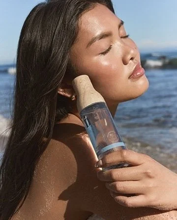 A woman at the beach holding a facial mist spray bottle near her face with eyes closed and a serene expression.