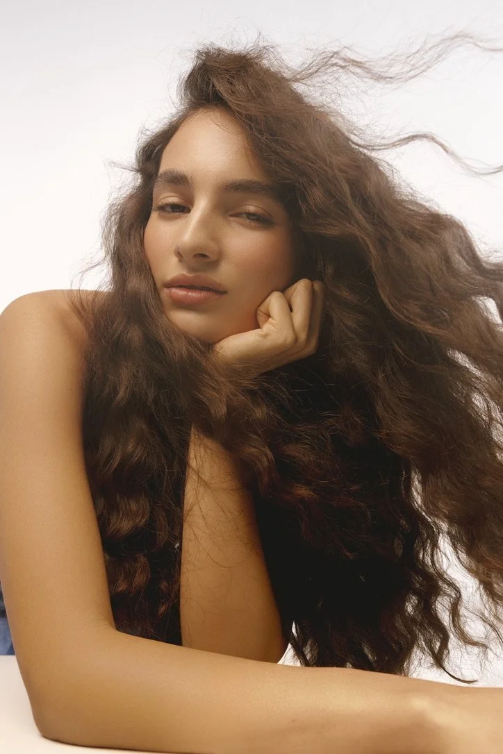 A woman with long, wavy brown hair posing with her chin resting on her hand, looking into the camera with a slight smile and one eye partially closed. Hair and makeup by Amber Rose.