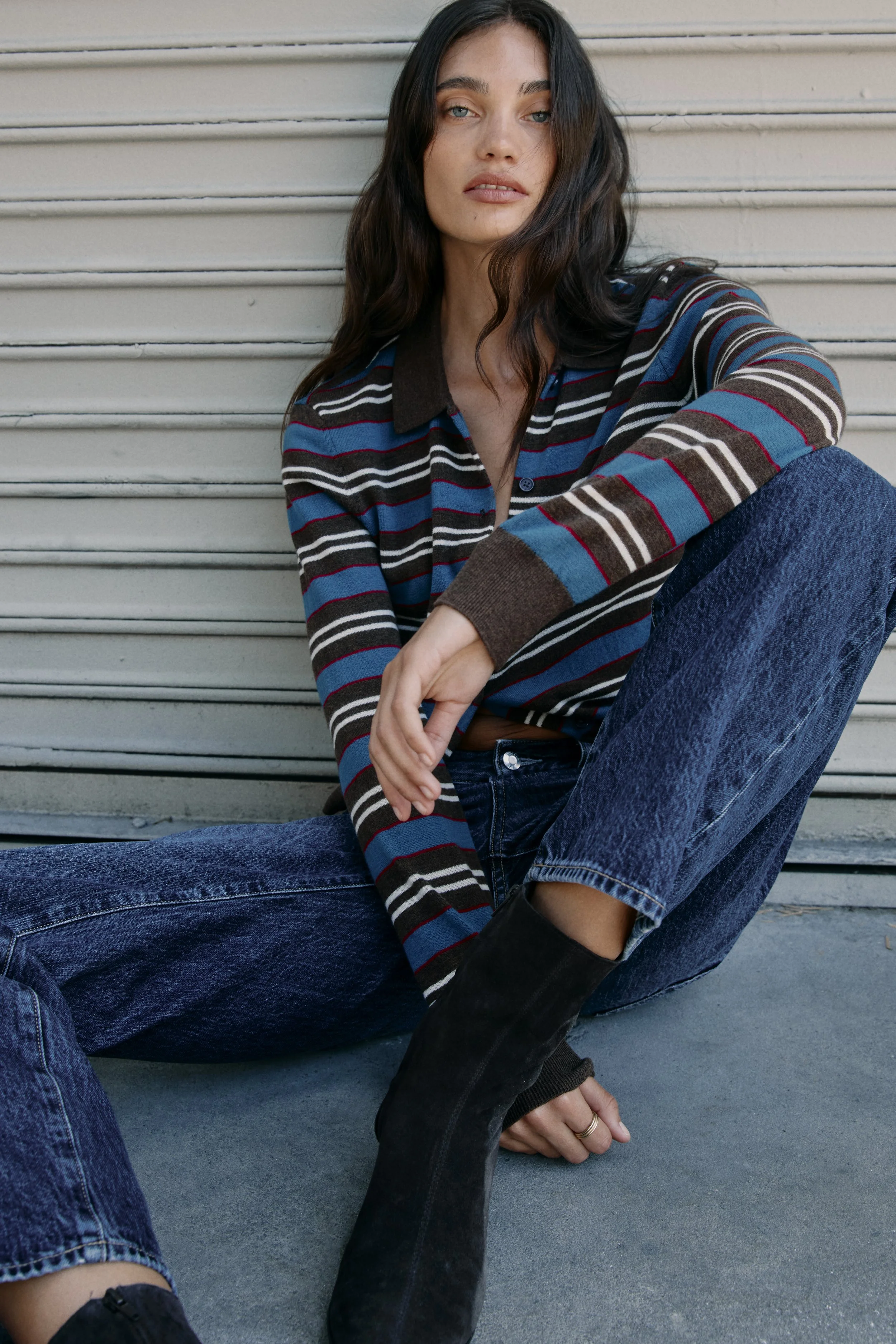 A young woman with long dark hair and blue eyes sitting against a gray wall with horizontal lines. She is wearing a striped shirt in shades of brown, blue, black, and white, paired with blue jeans and black boots. She has a relaxed pose with her righ
