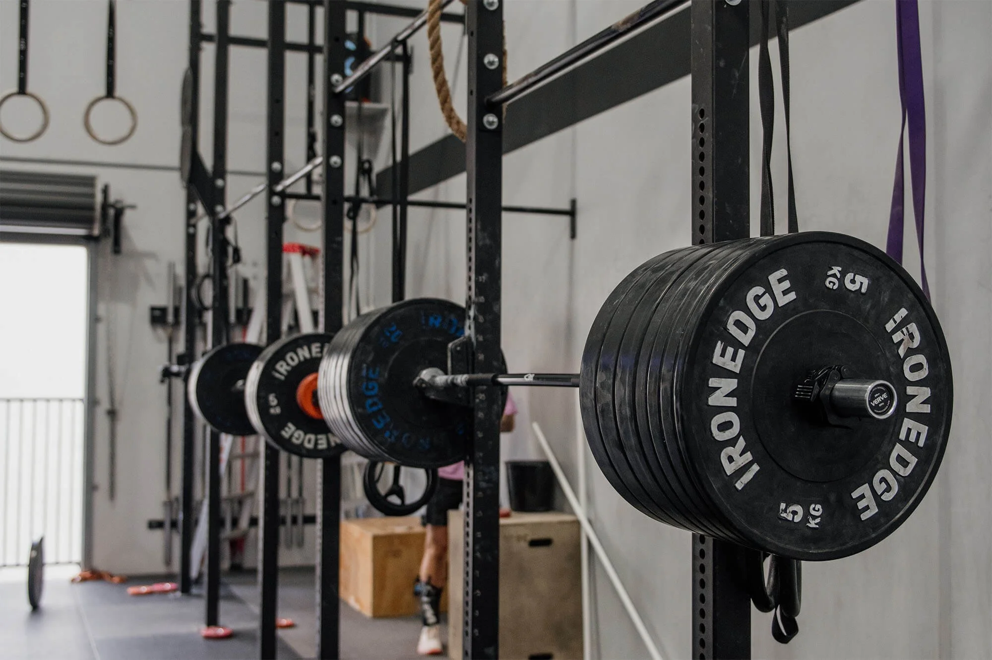 A photo at CrossFit TARIS of a barbell racked on the gymnastics rig in the foreground, and rings, plyo boxes and barbells in the background.