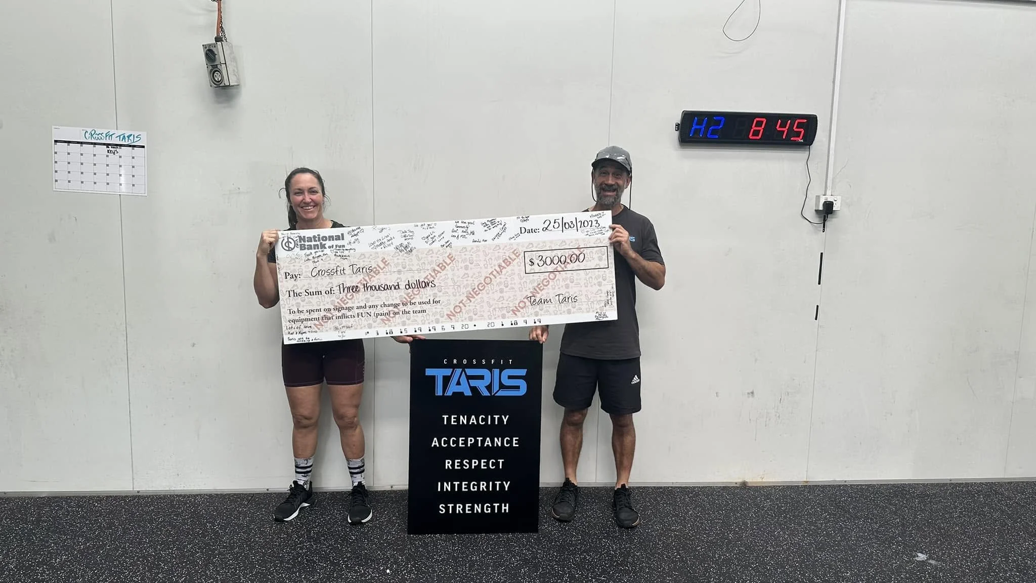 Affiliate owners Lisa and Shane Lake posing with an oversized pretend cheque from their members to donate towards their gym, CrossFit TARIS. In front of them is a sign with the meaning of TARIS - tenacity, acceptance, respect, integrity and strength.