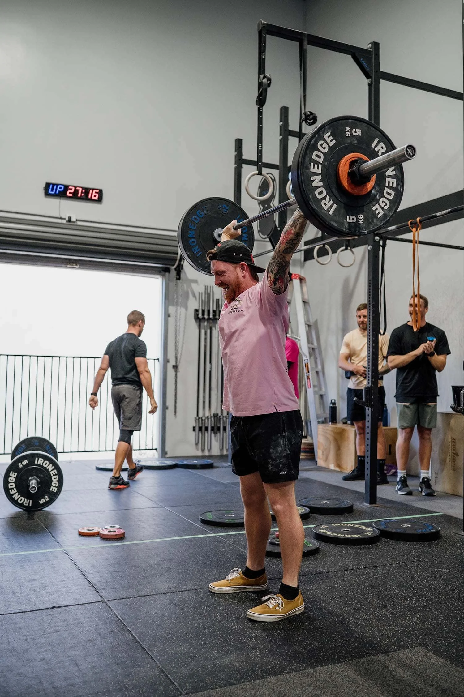 A male member completing a snatch during CrossFit TARIS's annual in-house weightlifting competition, with other members cheering in the background.