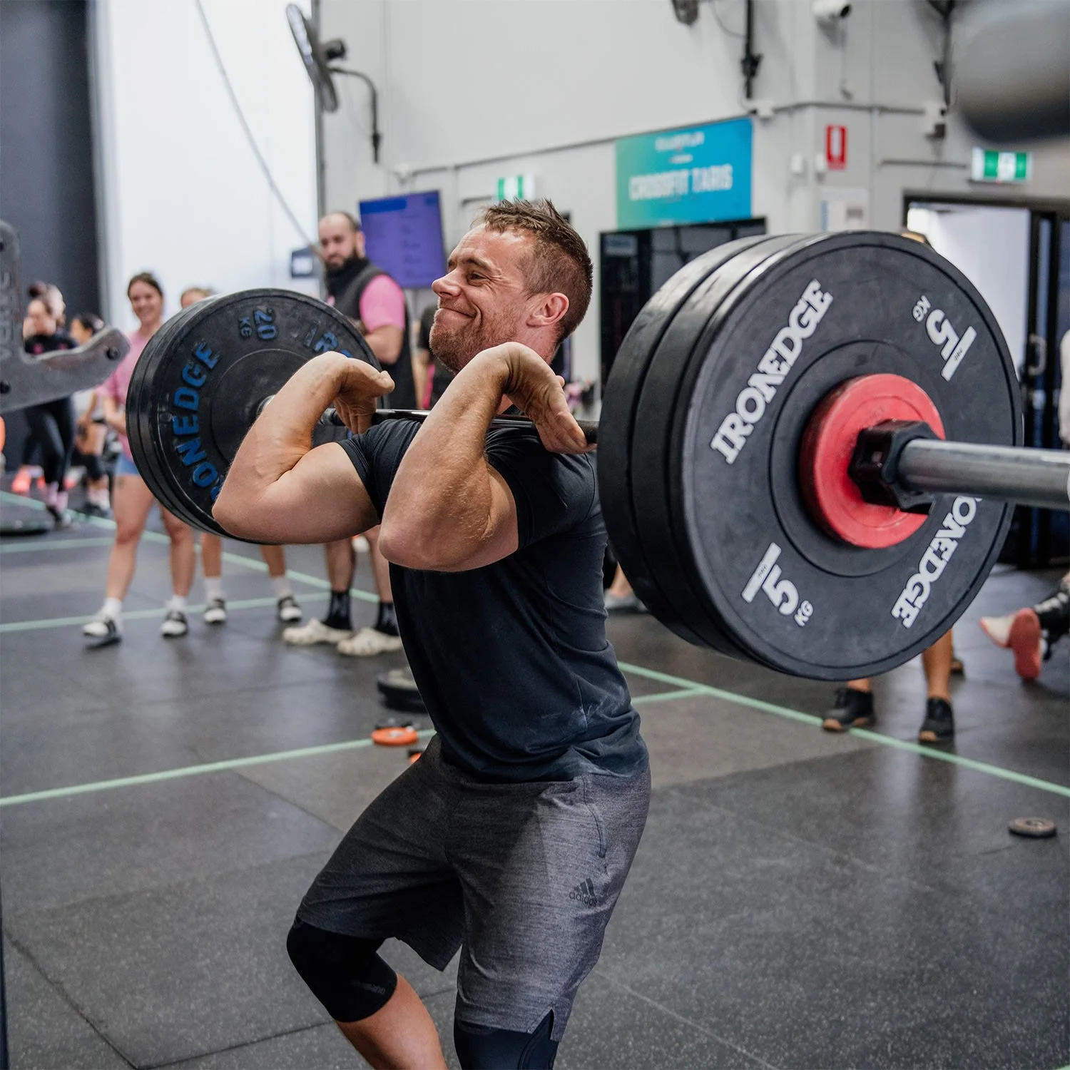 A male athlete at CrossFit TARIS in Logan completing a heavy front squat, with other members cheering him in the background.