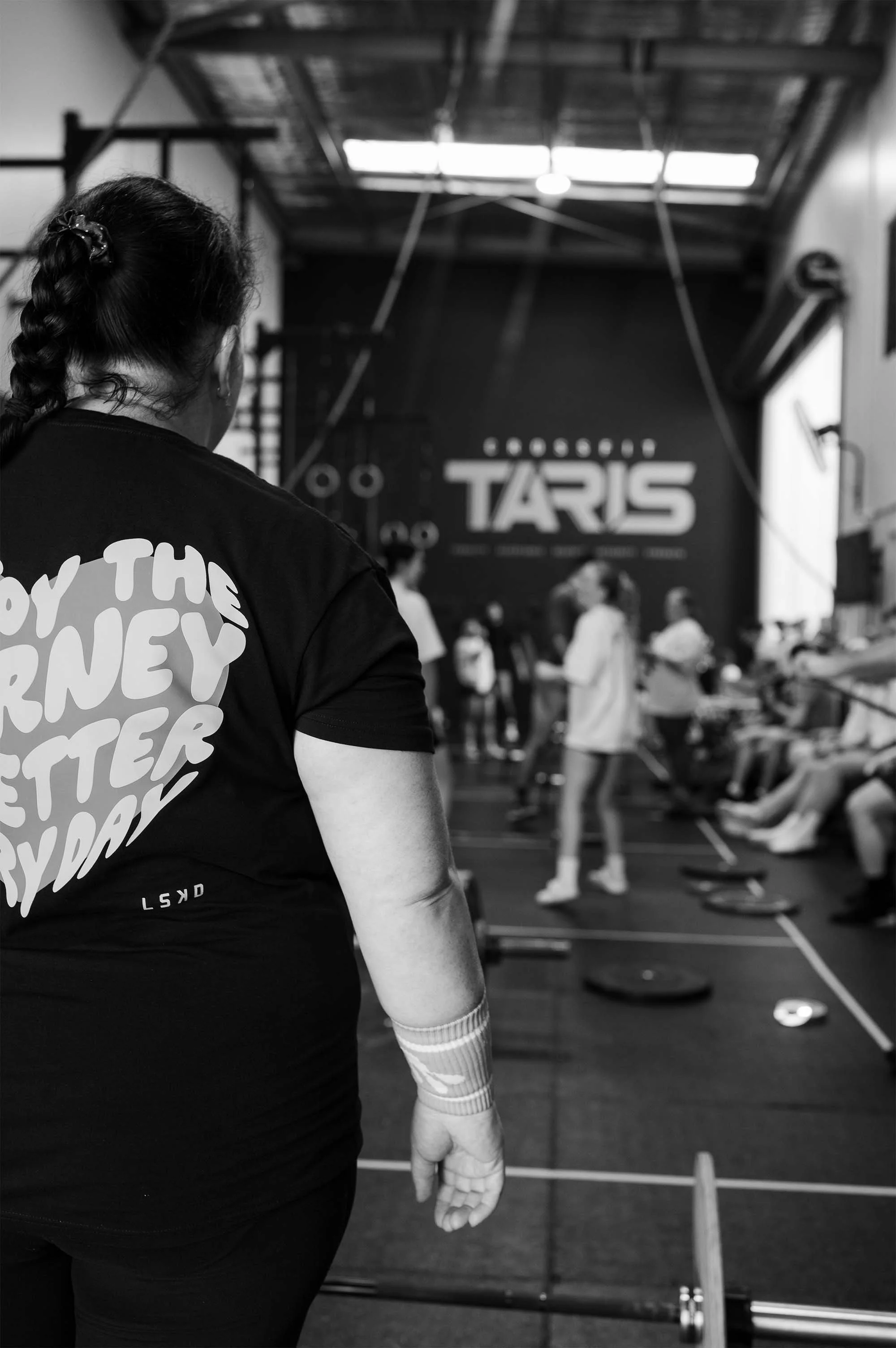 A female member of CrossFit TARIS wearing a LSKD shirt with her back to the camera, and other members blurred out in the background socialising.