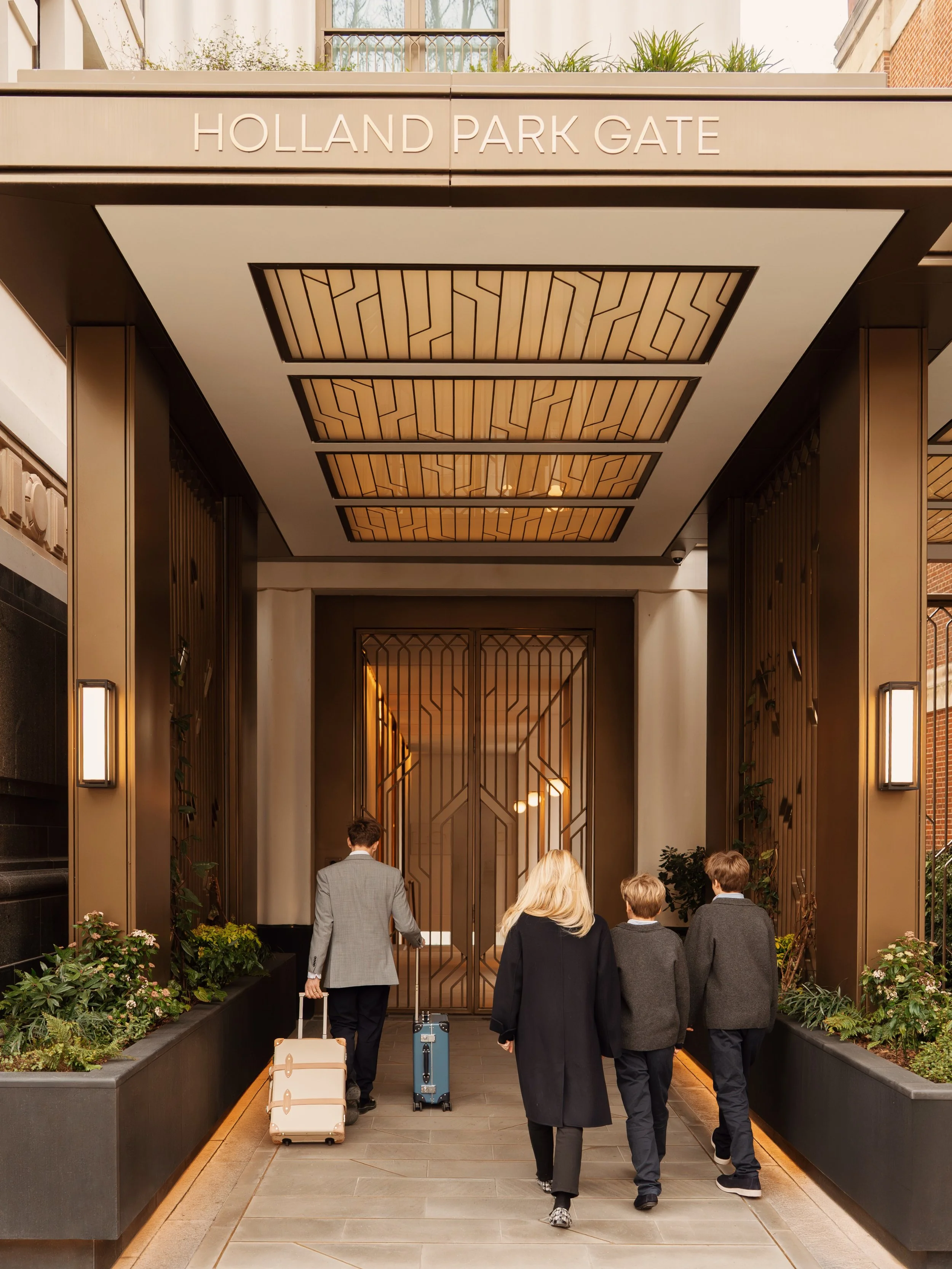Porter escorting a family with luggage through the planted pedestrian entrance at Holland Park Gate, Kensington, London