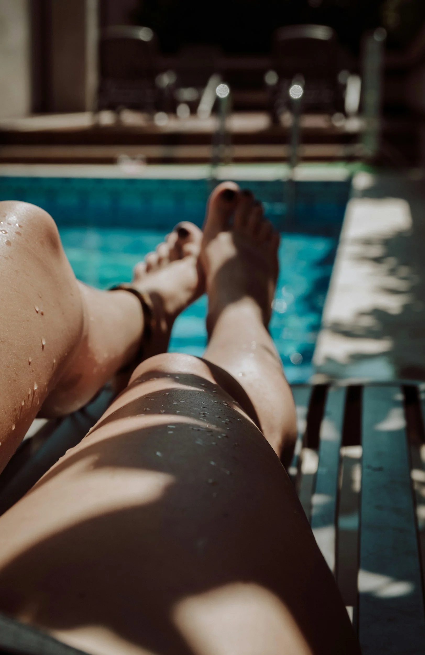 Personne allongée au bord de la piscine, jambes croisées, en train de se détendre sous le soleil, avec des chaises longues en arrière-plan.