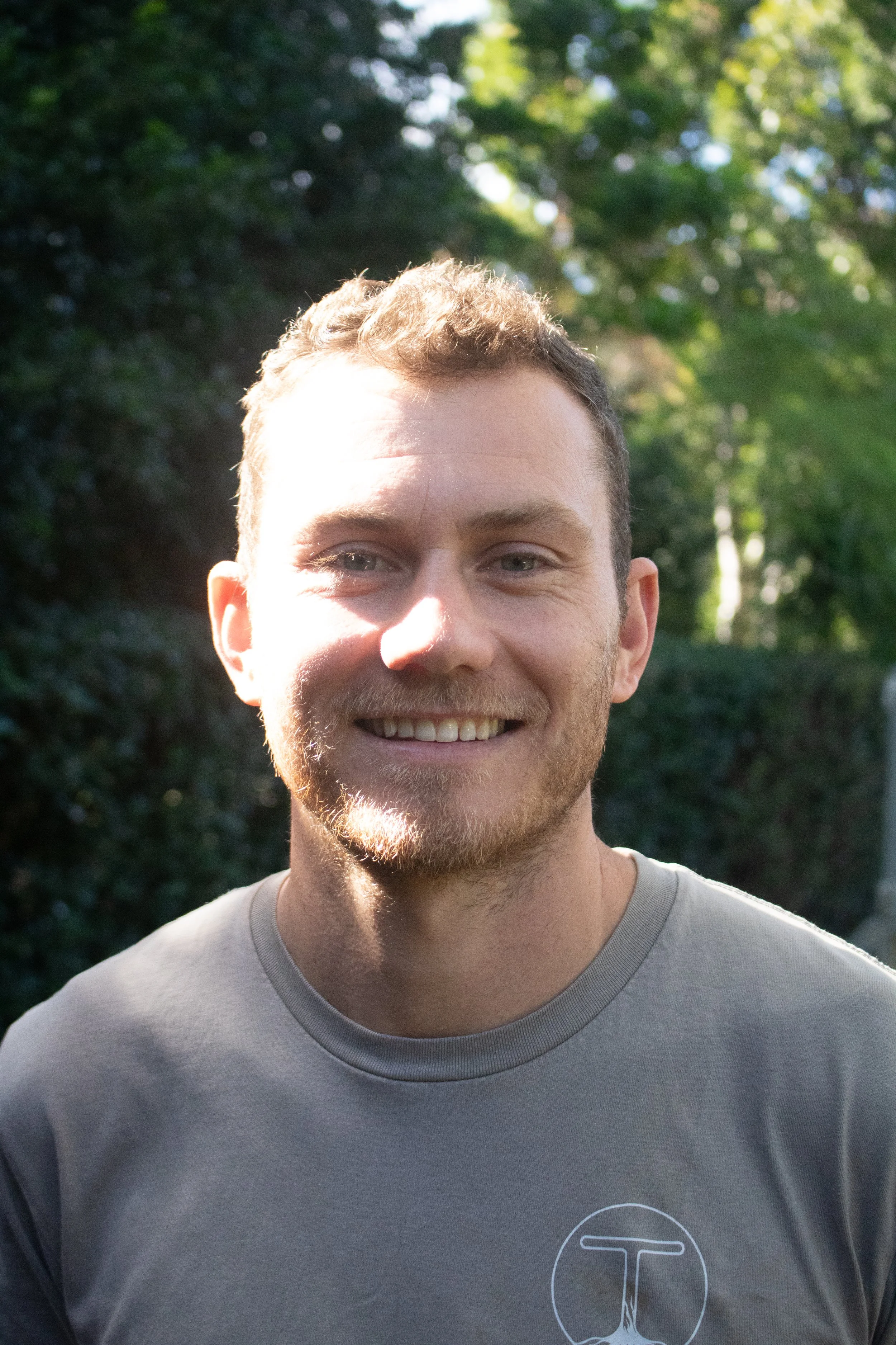 Close-up of a smiling man with curly hair and a beard, wearing a light gray T-shirt with a logo, outdoors with green trees in the background.