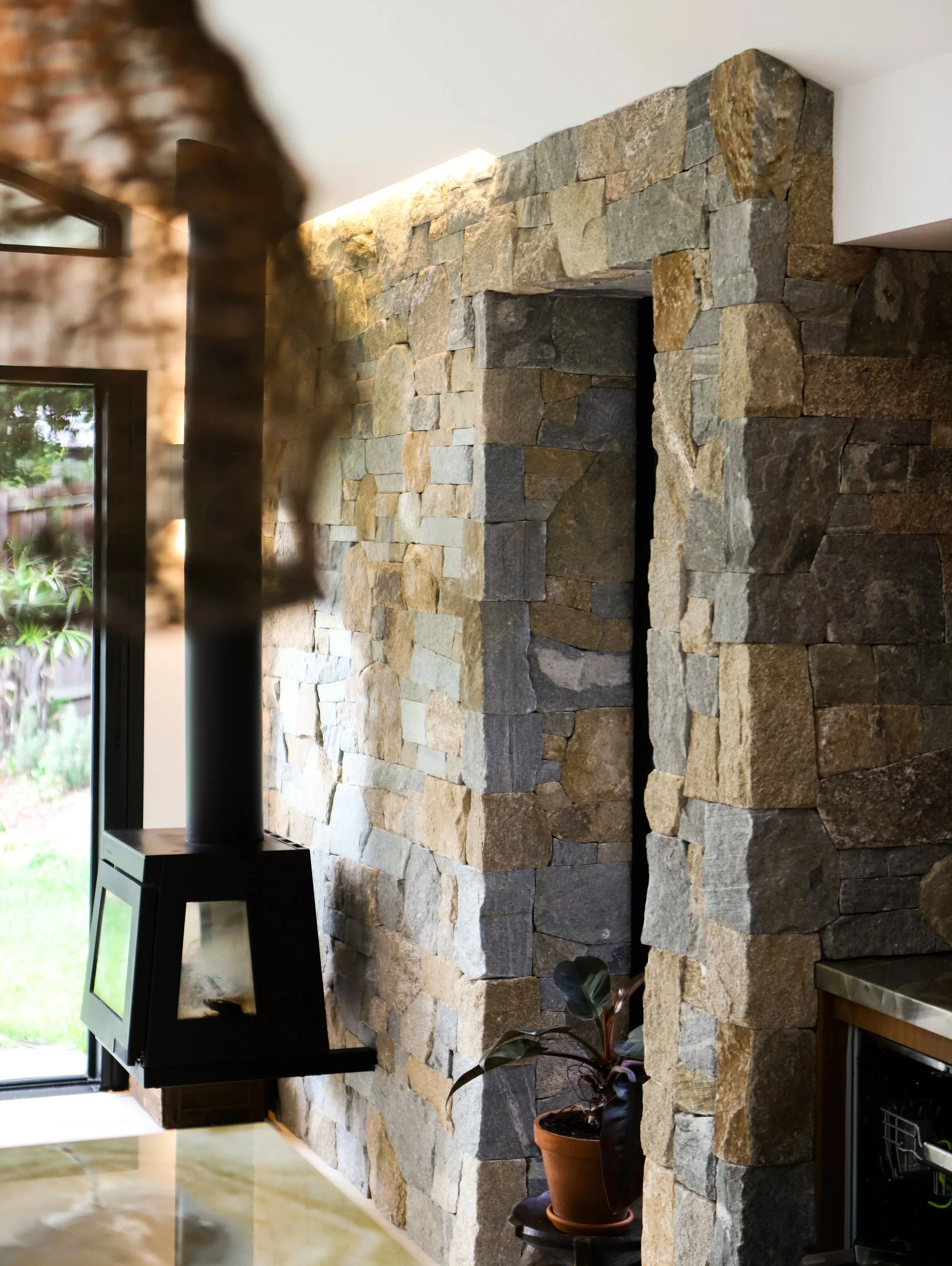 Interior view of a stone wall with a black wood-burning stove, a potted plant, and a window showing outdoor greenery.
