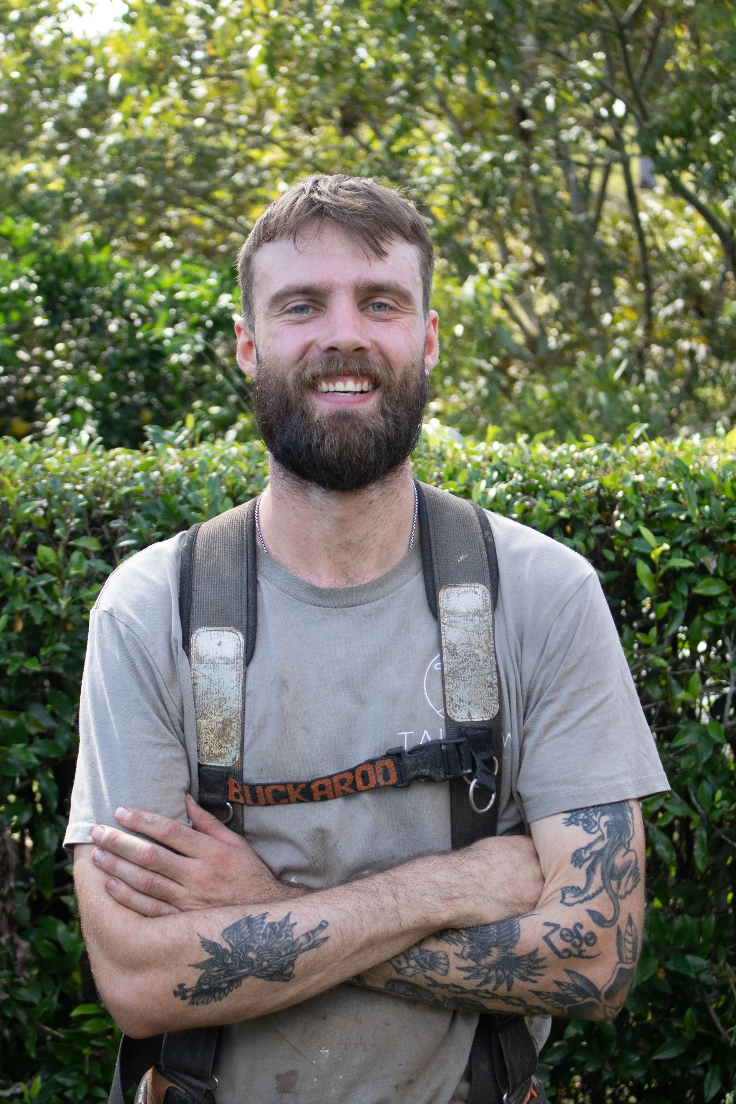 A smiling man with a beard and tattoos on his arms, wearing a gray t-shirt and a backpack with the brand Buck-Ard, standing outdoors in front of greenery.