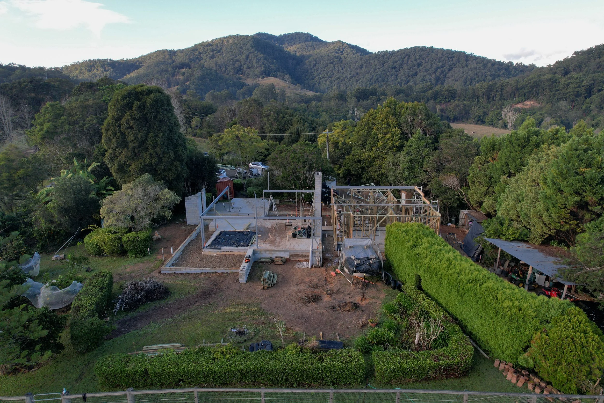 Aerial view of a house under construction in a green, hilly landscape with trees and mountains in the background.