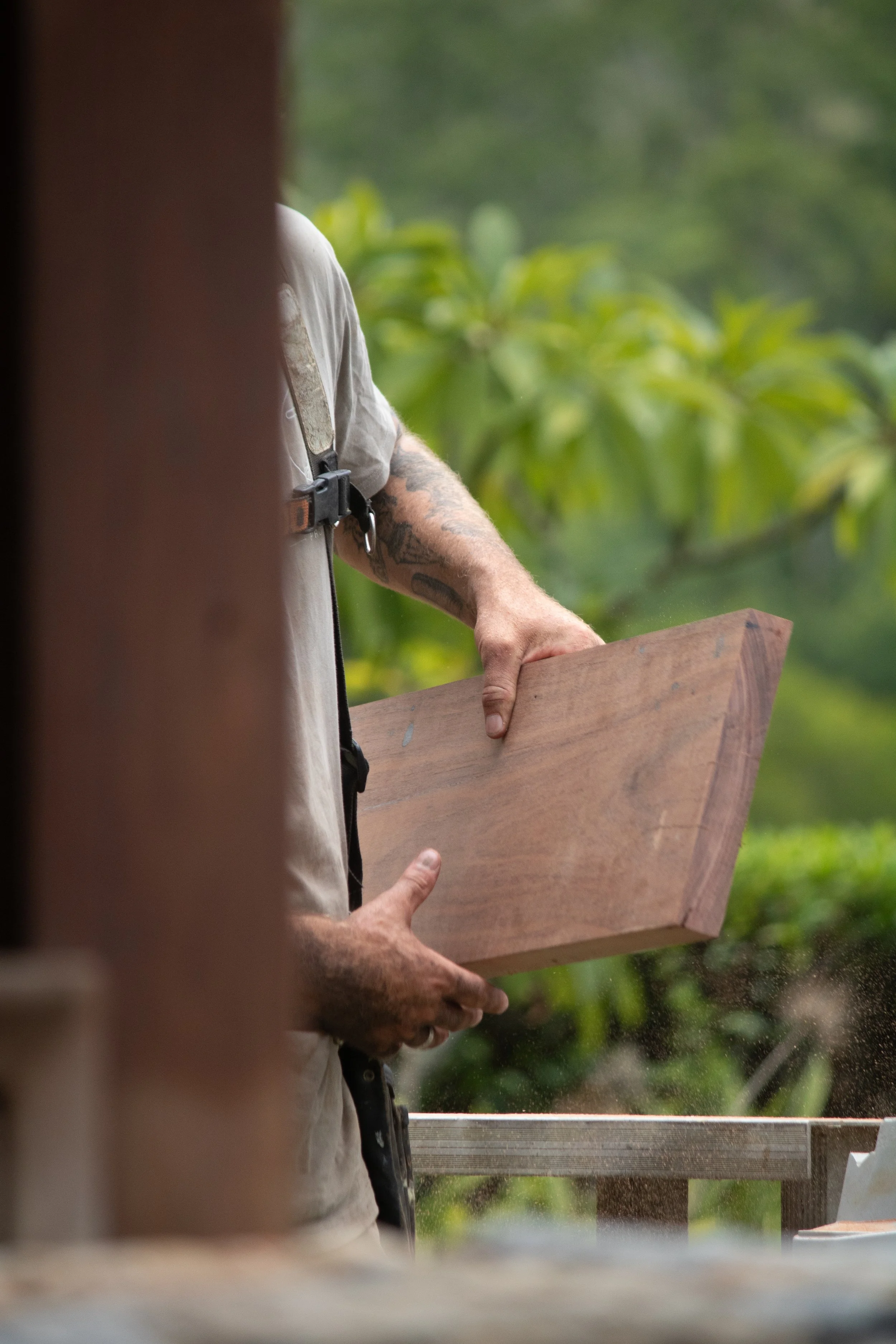 A person wearing a white t-shirt holding a wooden plank outdoors with green foliage in the background, partially obscured by a vertical wooden beam or structure.