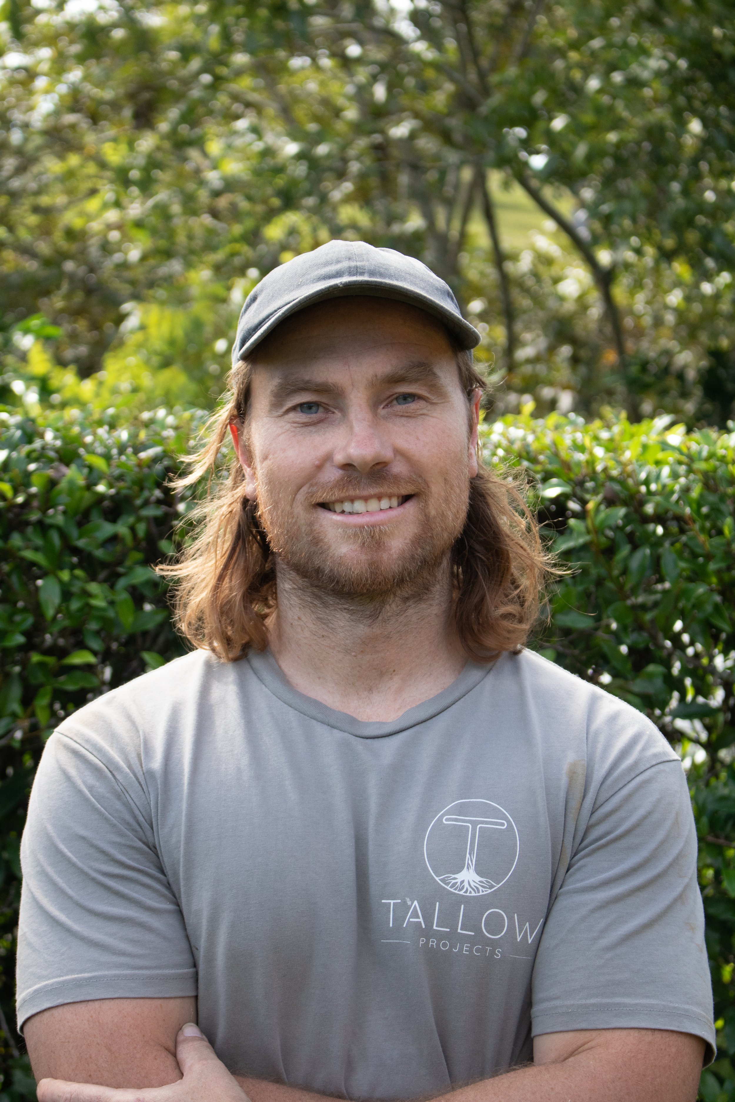 A man with long reddish-brown hair and a beard is standing outdoors in front of a green bush with trees in the background. He is wearing a light gray T-shirt with a logo and text that says 'Tallow Projects' and a gray baseball cap. The man is smiling and looking directly at the camera with his arms crossed.