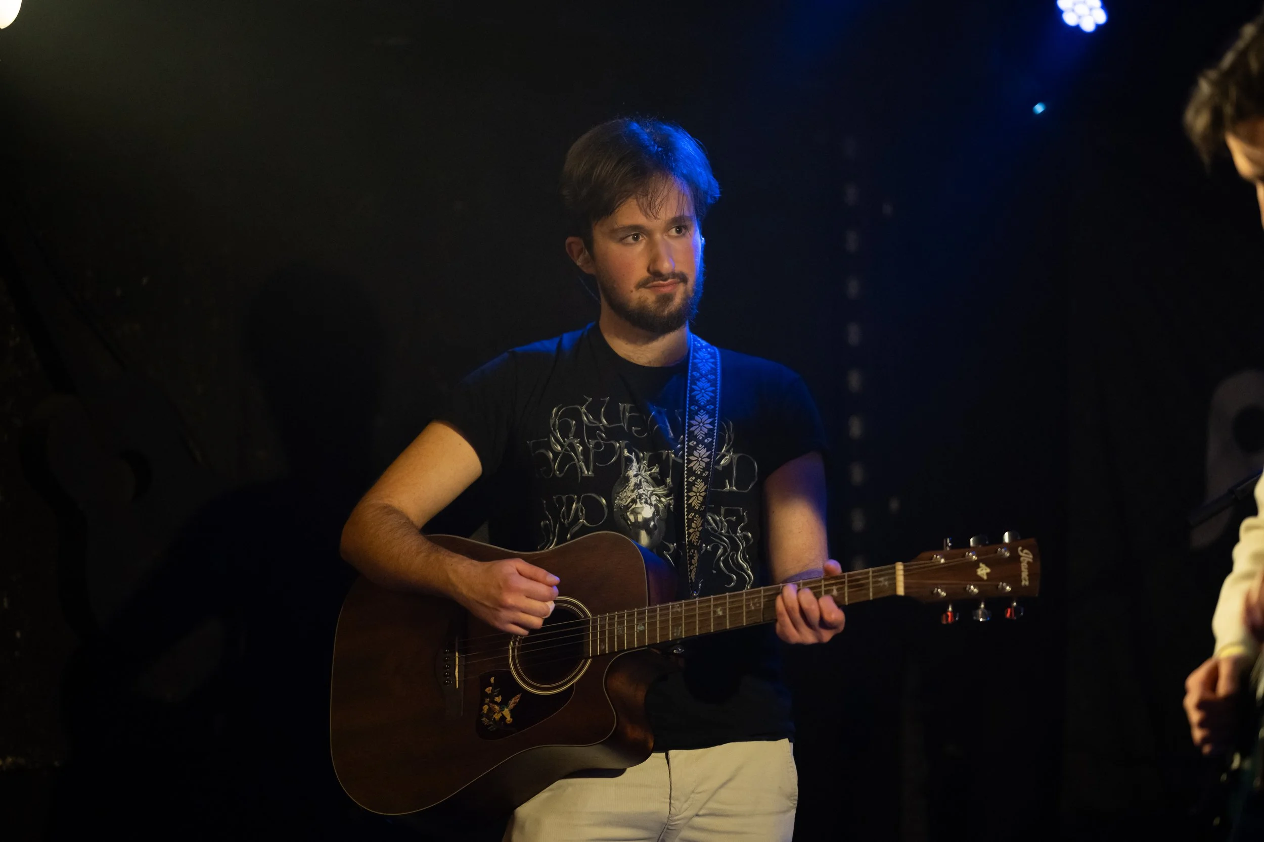 A young man with dark hair and a beard playing an acoustic guitar on stage, wearing a black T-shirt with a graphic design.