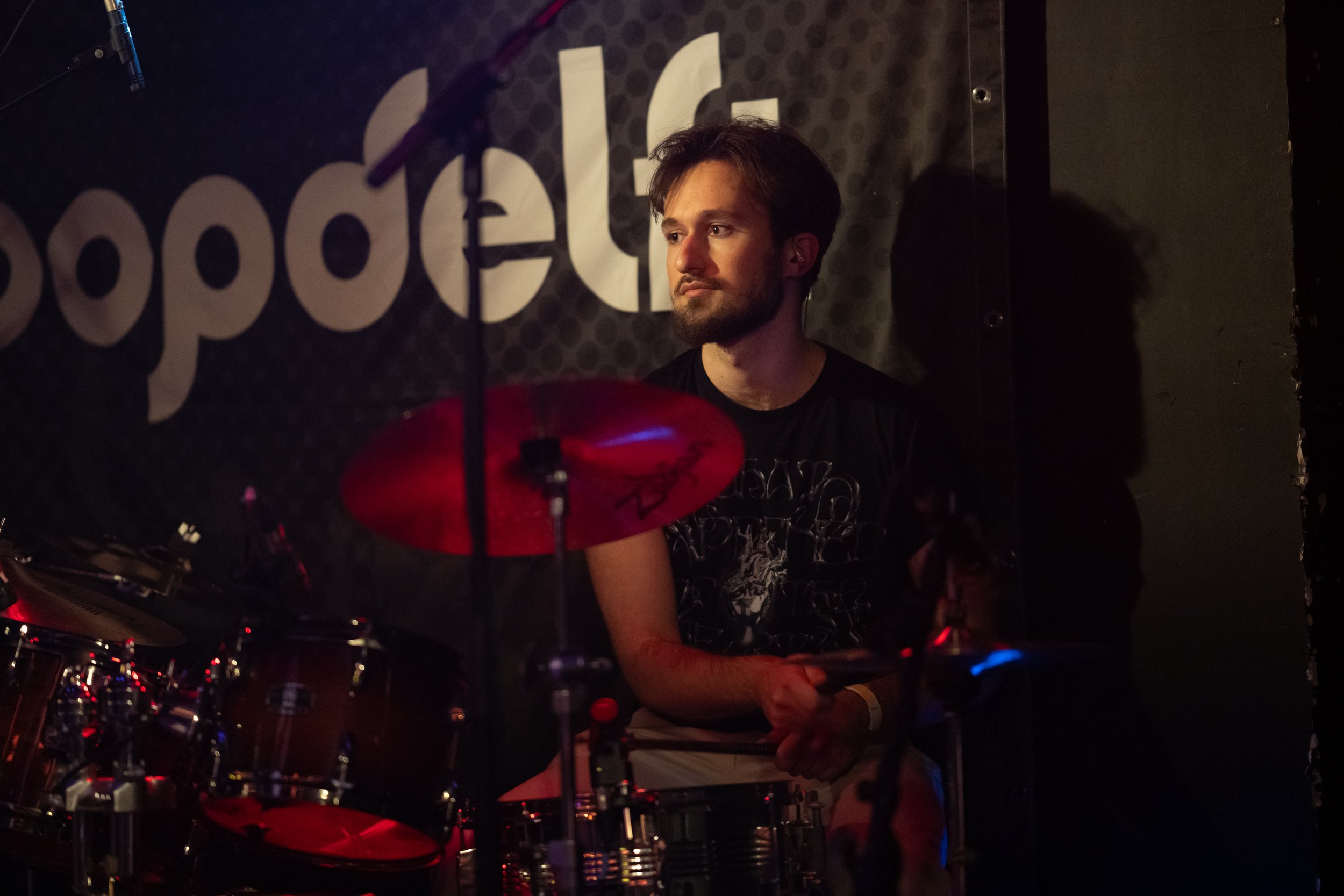 A man with dark hair and a beard playing drums on stage at a music venue. The stage has a black background with large white letters that spell out 'popPlace'. There is a microphone hanging from above, and the image is dimly lit with some red and blue