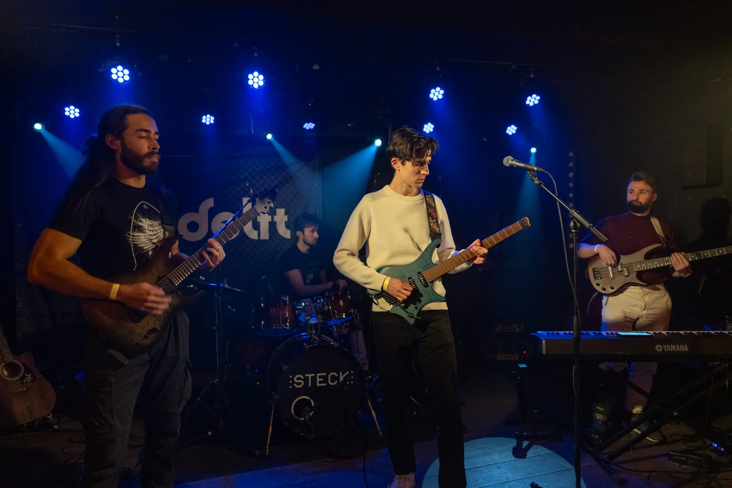 A band of four young men performing on stage in a dimly lit venue with blue stage lighting. Two of them are playing electric guitars, one is playing bass, and one is behind a drum set, with a keyboard in the foreground.