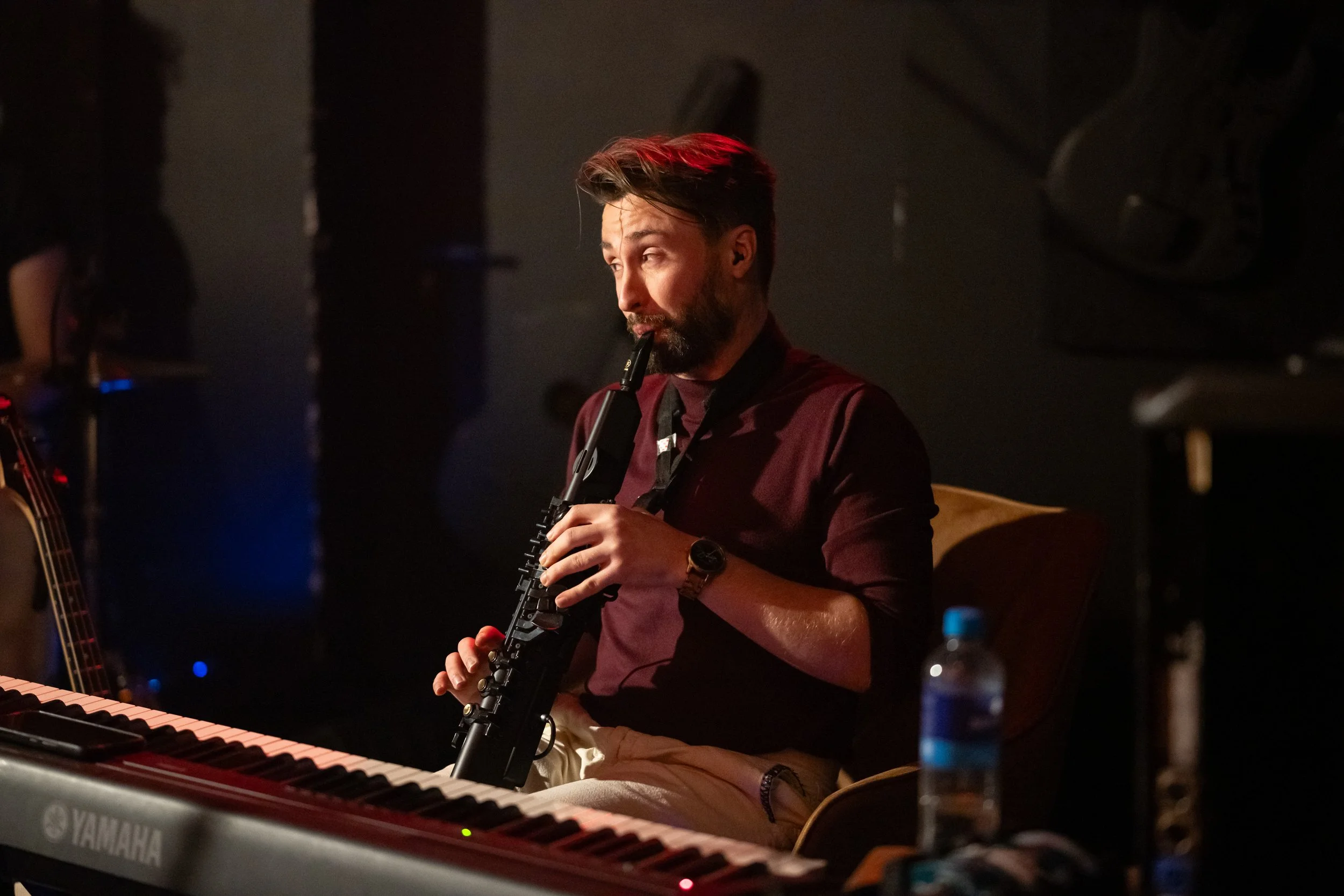 A man with dark hair and a beard playing a melodica in a dimly lit setting, with a Yamaha keyboard in the foreground.