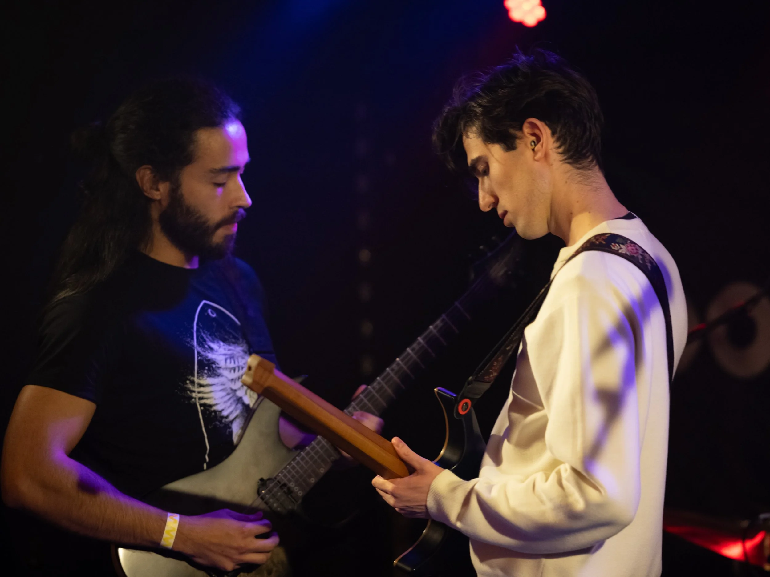 Two young men with dark hair and facial hair playing electric guitars in a darkened room with purple and red stage lighting.
