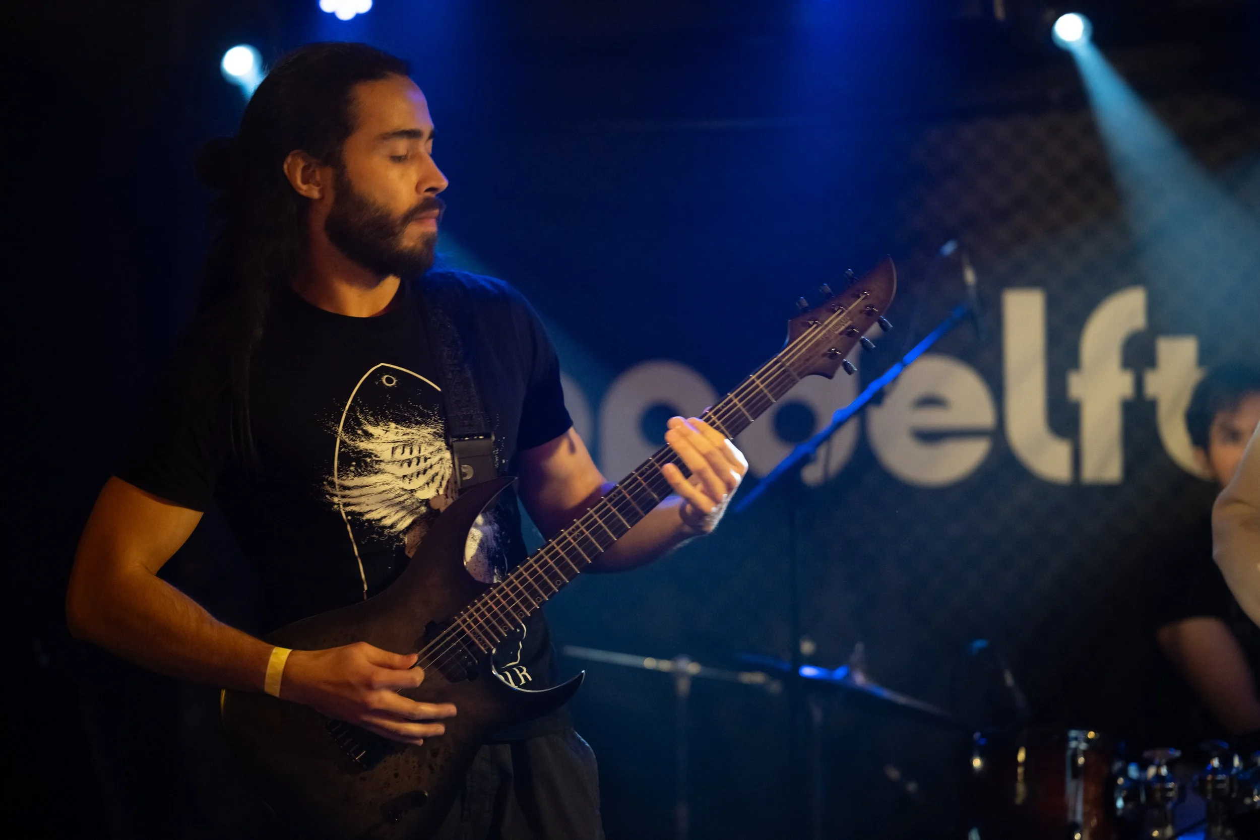 A man with long dark hair tied back, a beard, and mustache playing an electric guitar on a stage with blue and white spotlights, and a large sign with partially visible text in the background.