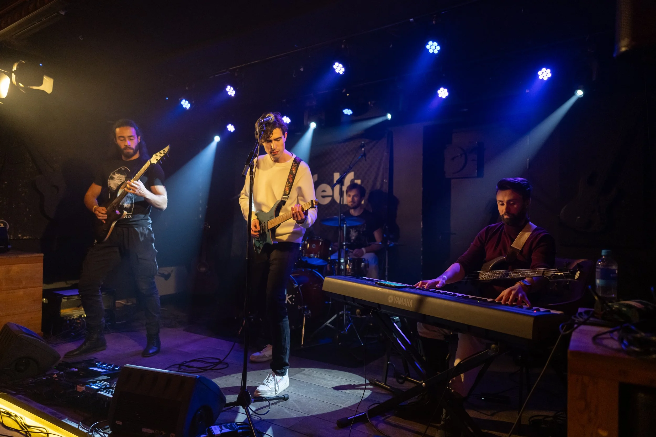 A band performing live at a music venue. Four musicians are visible: one playing bass guitar, one playing electric guitar, a drummer in the background, and a keyboardist in the foreground. The stage is dimly lit with blue and white spotlights.