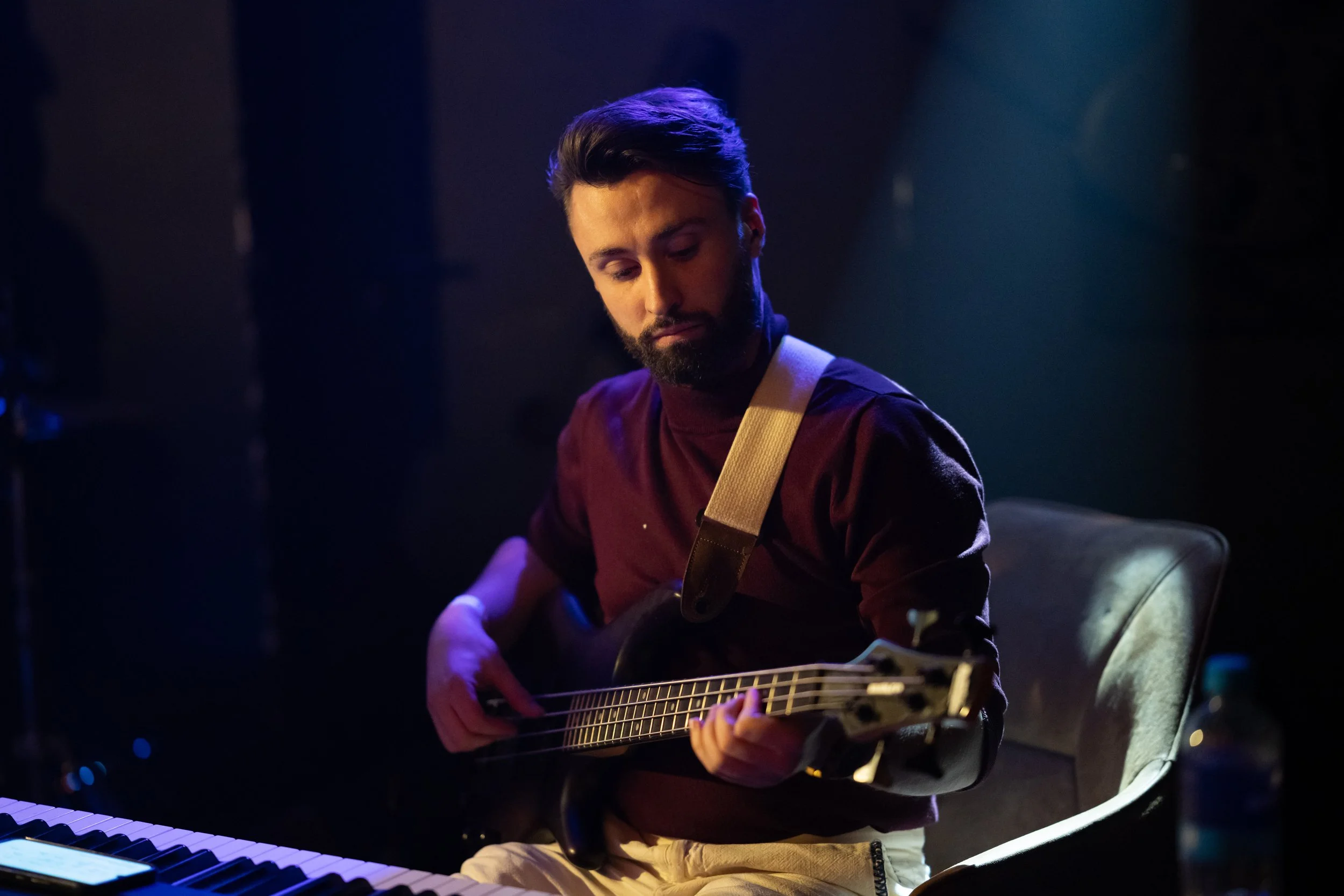 A man with dark hair and a beard wearing a maroon shirt and beige pants plays an acoustic guitar while sitting in a dimly lit room with blue and purple lighting.