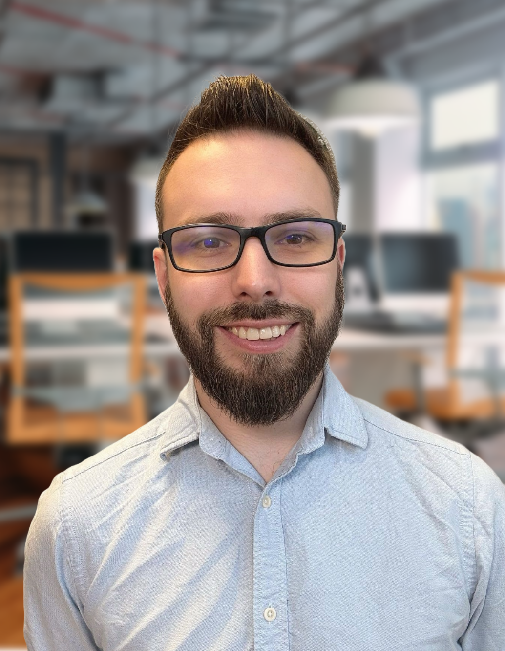 A man with glasses, a beard, and short hair smiles for a photo in an office space with desks and chairs in the background.
