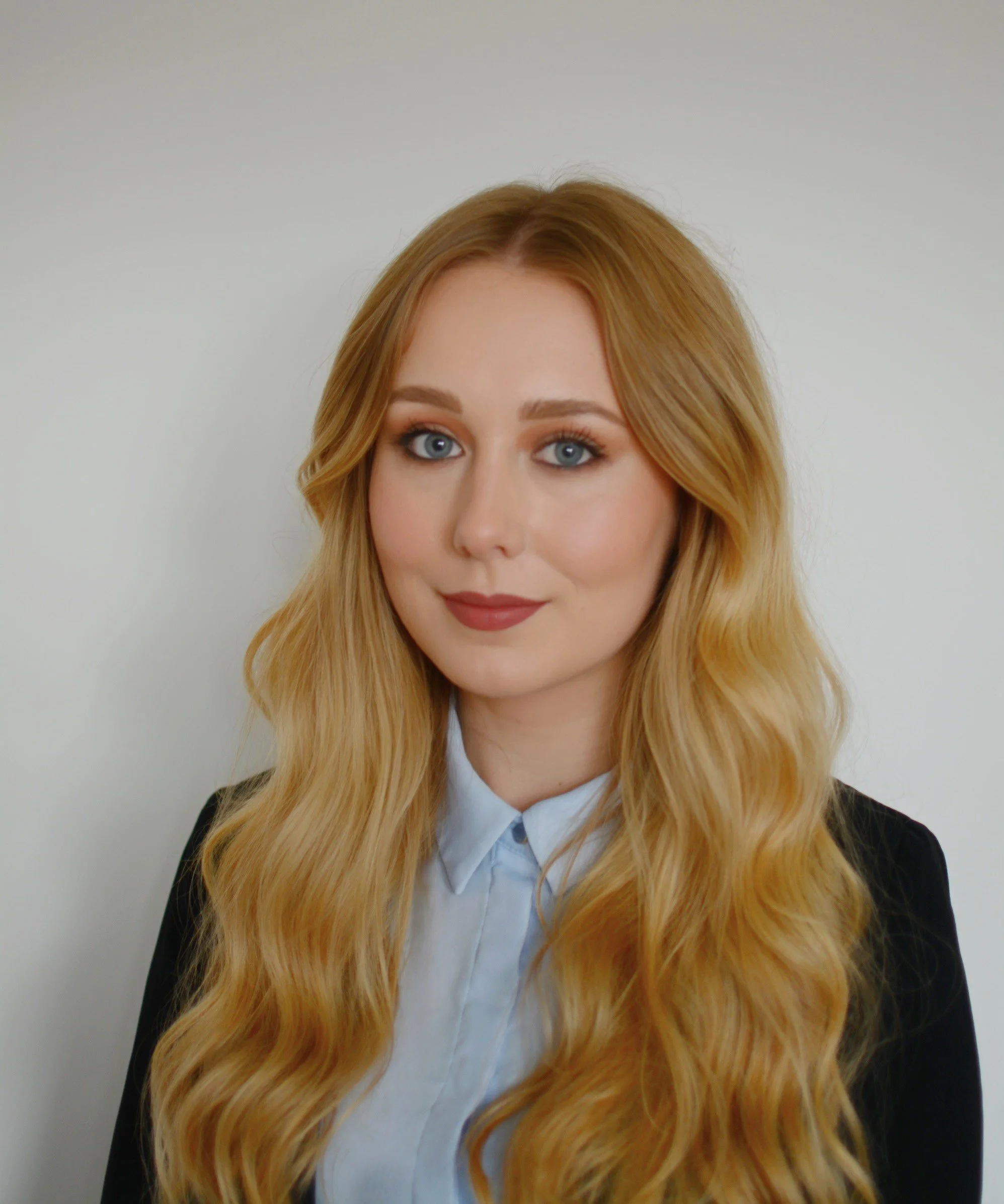 A young woman with long, wavy blonde hair, blue eyes, wearing a light blue collared shirt and black blazer, posing against a plain white background.