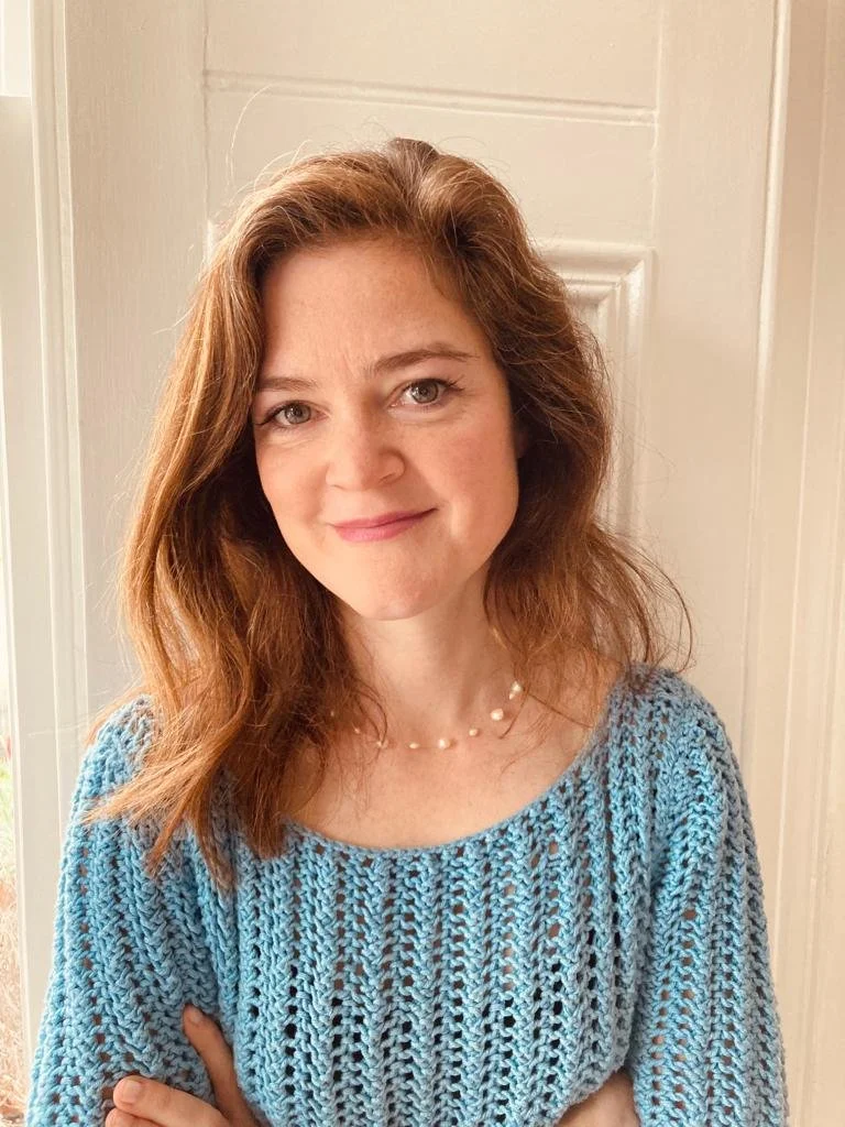 A woman with shoulder-length wavy red hair, wearing a light blue crochet top and a pearl necklace, standing in front of a white door, smiling.