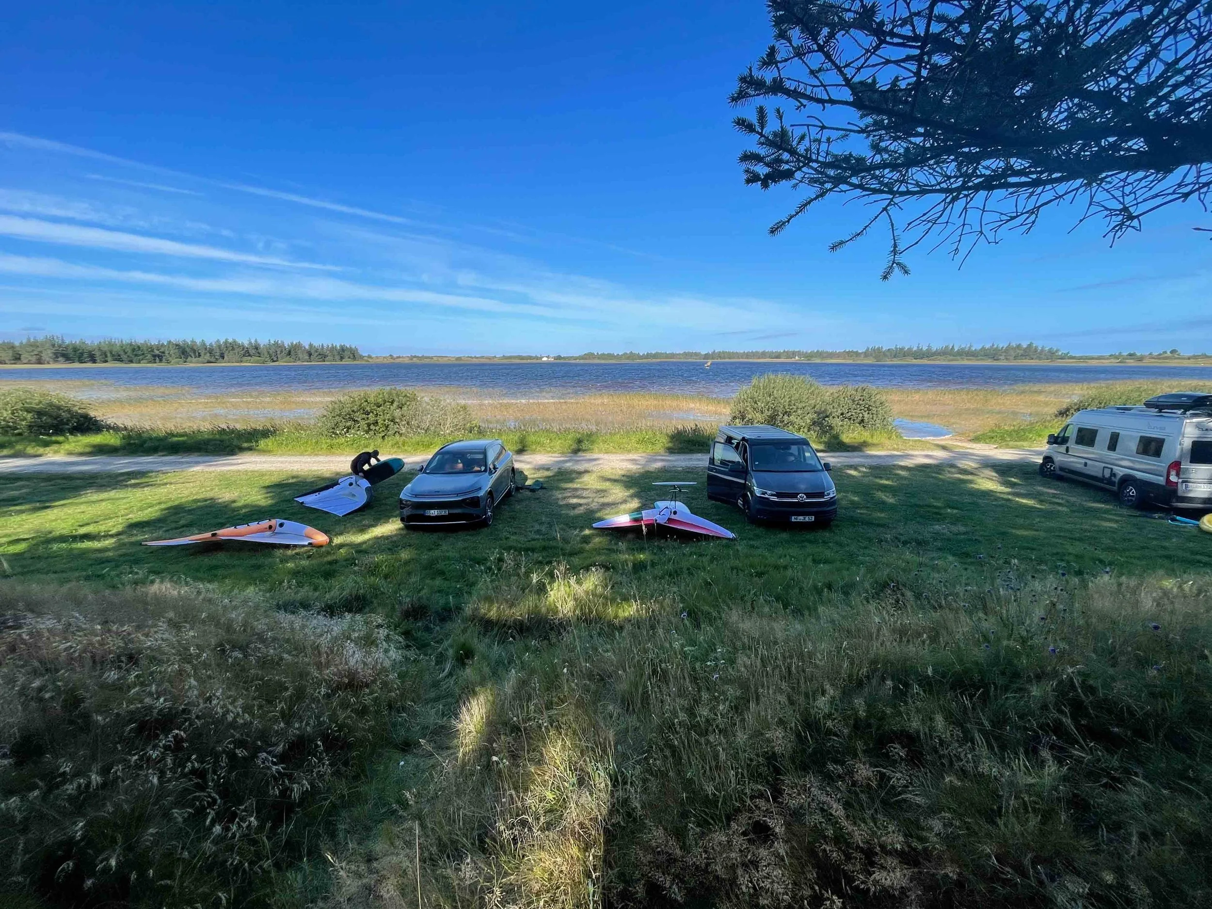 Ein Park am Wasser, mit zwei Autos, mehreren aufblasbaren Kajaks und einem Rucksack, im Schatten eines Baumes.