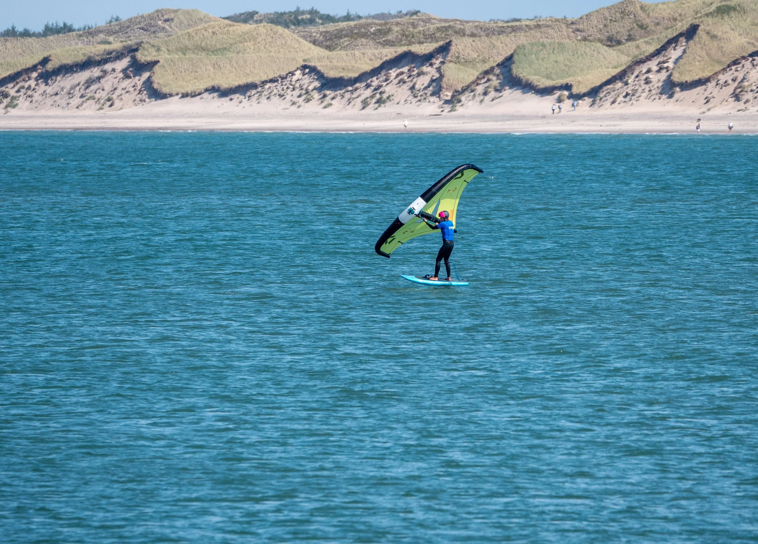 Menschen beim Wassersport auf dem Meer, mit einem Kitesurfer im Vordergrund, der eine große grüne und schwarze Kite segelt, im Hintergrund eine Küstenlinie mit Sandstrand und Dünen unter blauem Himmel.