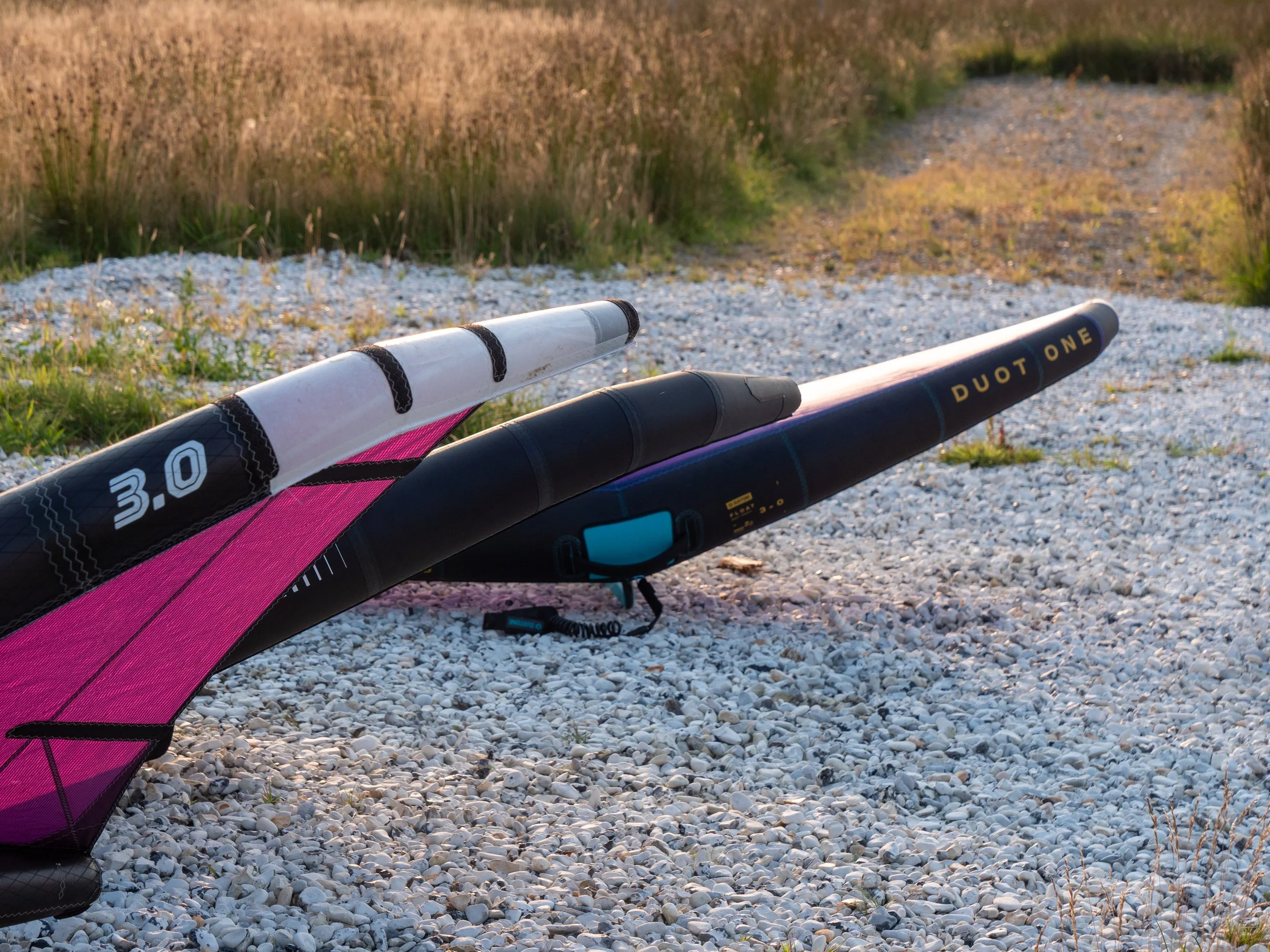 Zwei aufblasbare Wassersportgeräte, ein Wind- oder Kitesurf-Board und eine Surf- oder SUP-Paddle, liegen auf einem Kiesweg in einer Naturumgebung mit Gräsern und Sträuchern.