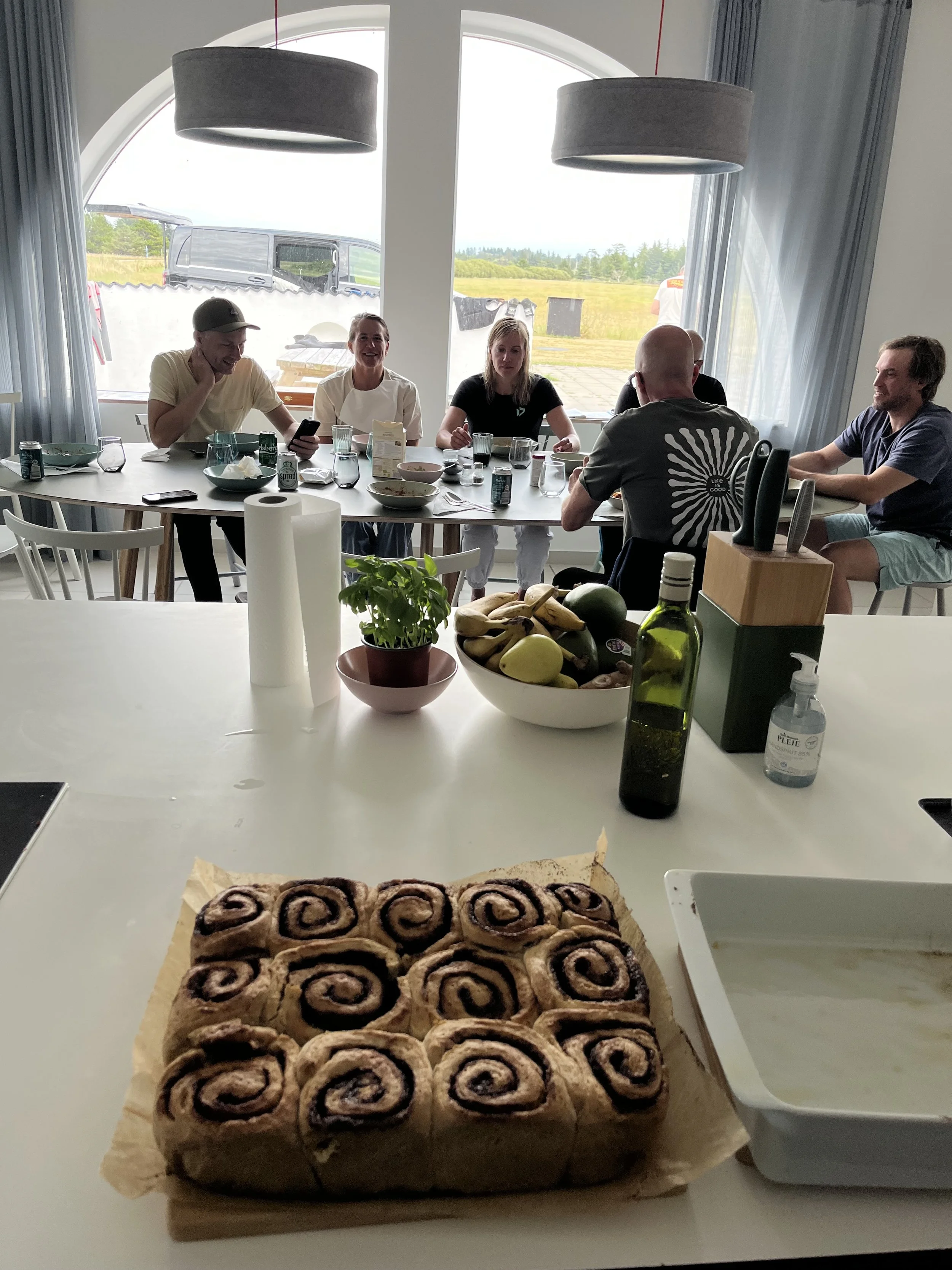 Draufsicht auf eine Küchenarbeitsfläche mit einer vorbereiteten Zimtschneckenrolle, frischem Basilikum, Bananen, einem Olivenölflasche und einem Papiertuchspender, im Hintergrund eine Mahlzeit beim Essen bei einem Treffen mit sechs Personen an einem 