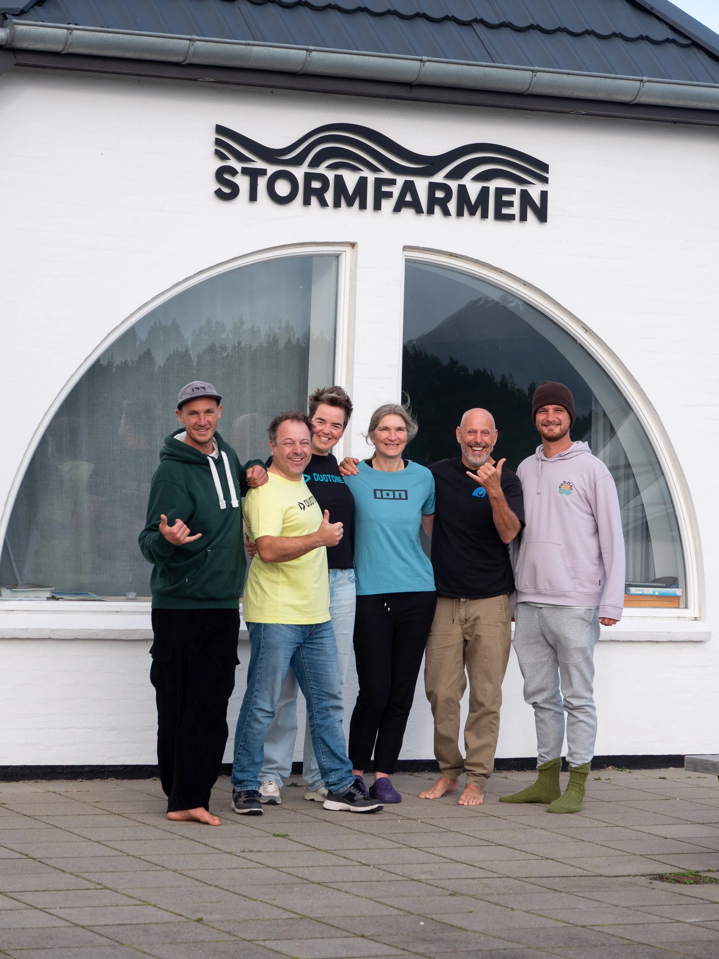 Sechs Menschen stehen vor einem weißen Gebäude mit einem schwarzen Wellen-Logo und der Aufschrift 'STORMFARMEN'.