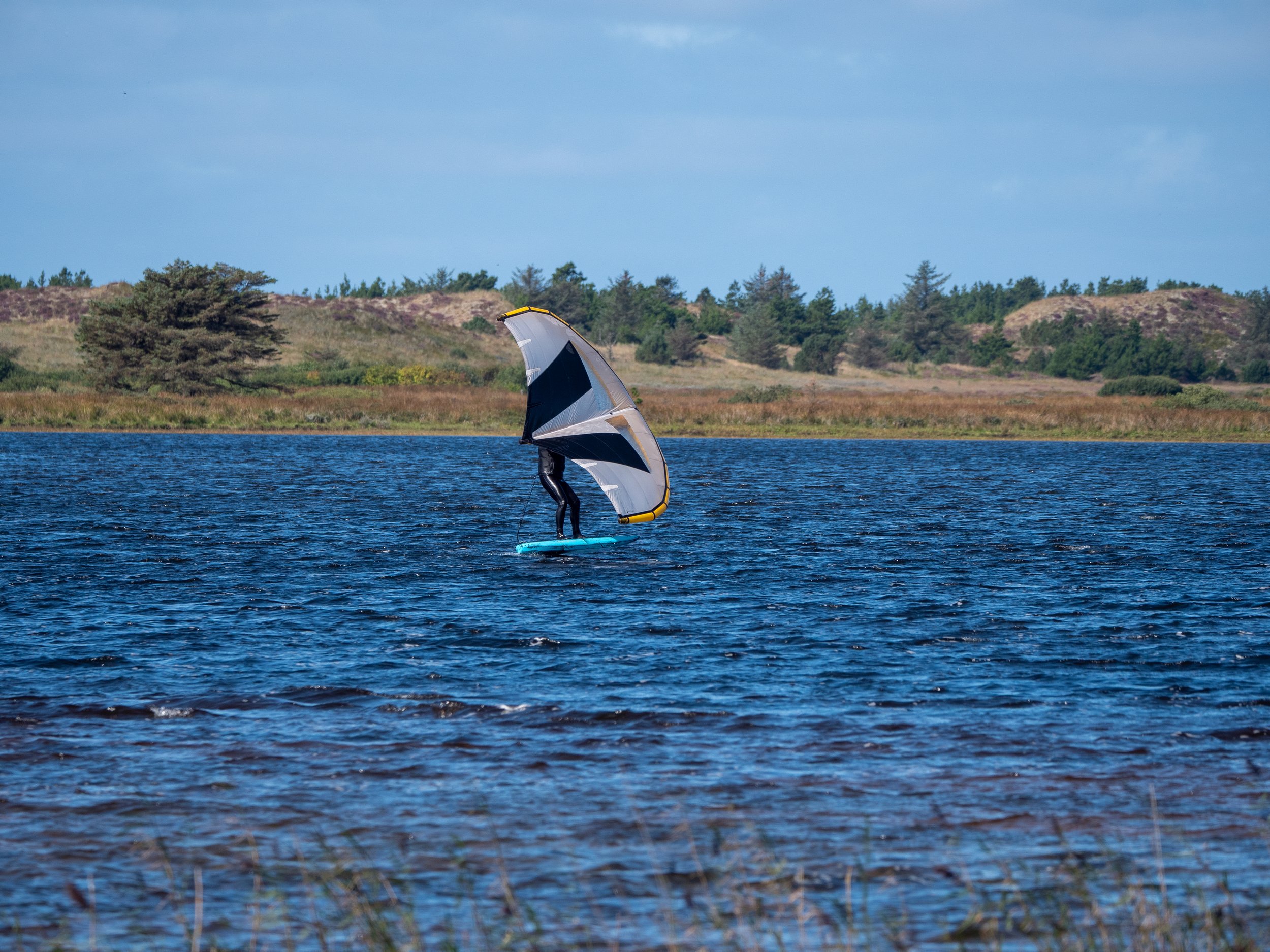 Ein Mensch steht auf einem Wasser-Surfbrett und benutzt ein große, aufgespannten Fallschirm, um sich über das Wasser zu ziehen, mit einer ländlichen Landschaft im Hintergrund.