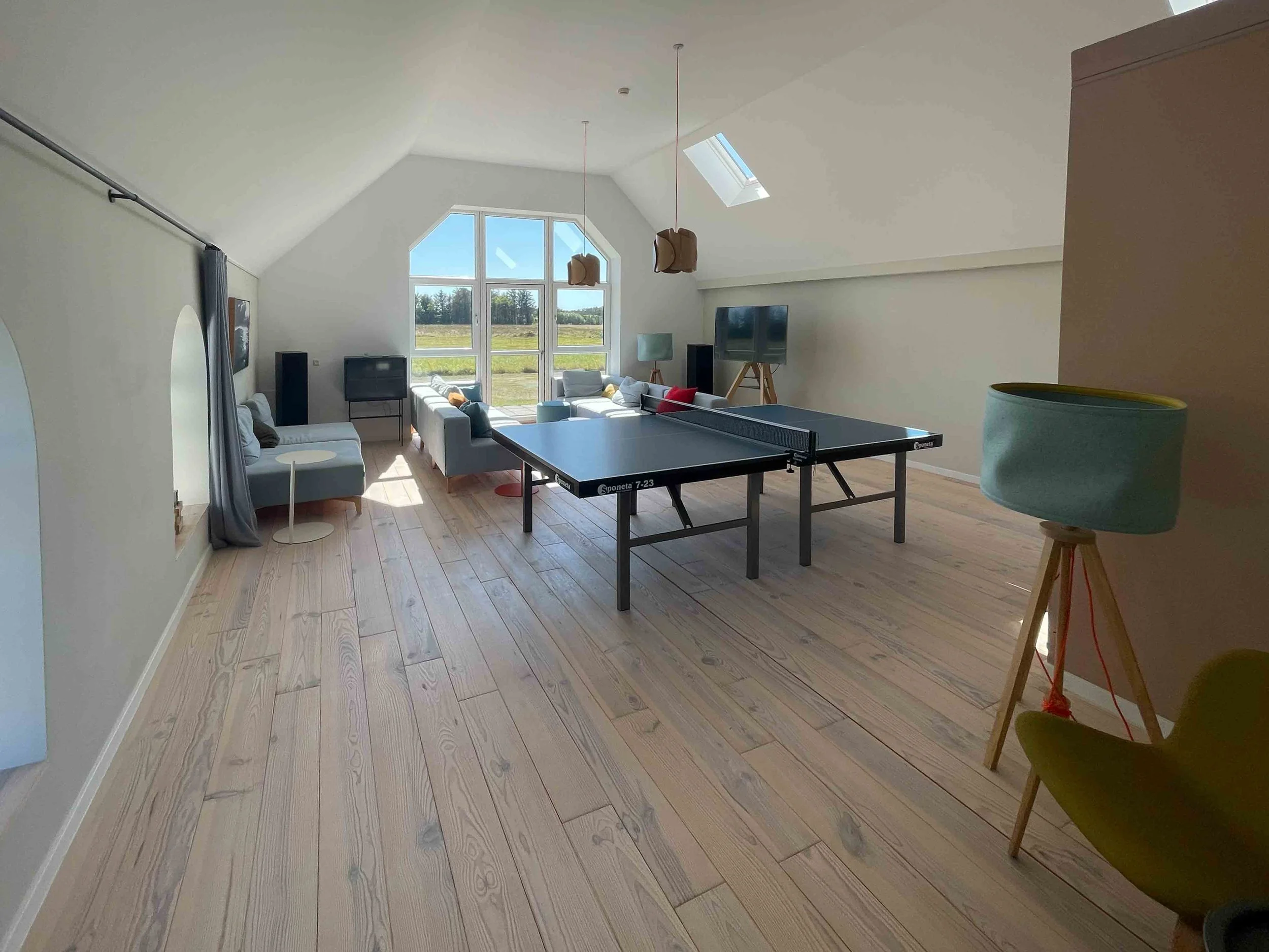 Ein heller, moderner Dachboden mit großem Fenster, auf dem sich ein Tischtennisschläger befindet, in einem gemütlichen Wohnzimmer mit Sofas, Fernseher und Stehlampen.