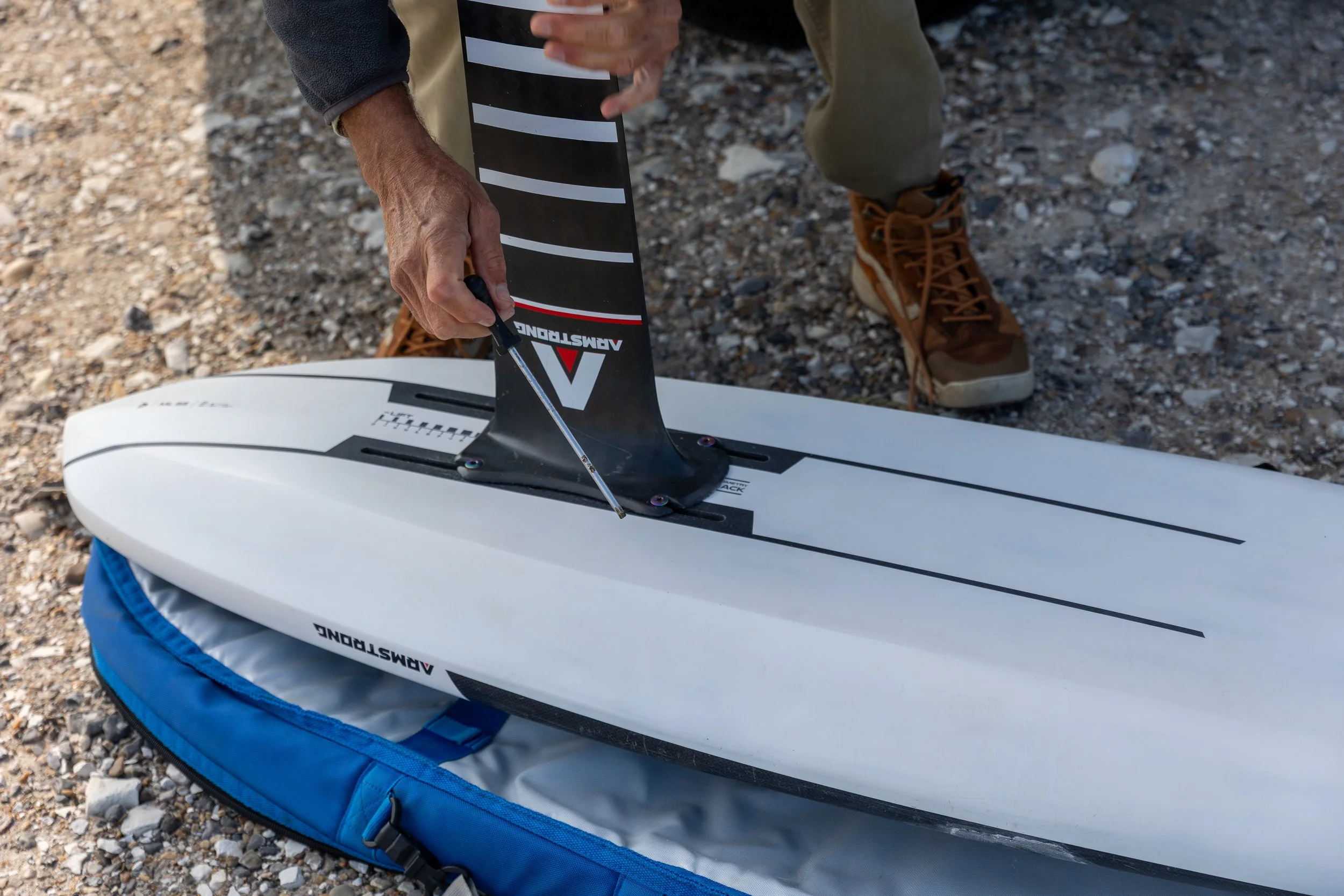 Eine Person bereitet eine Windsurf-Finne vor, indem sie sie am Deck eines Windsurf-Boards befestigt. Das Board liegt auf einer blauen Tragetasche auf einem Kiesboden, die Person trägt braune Schuhe und beige Hosen.