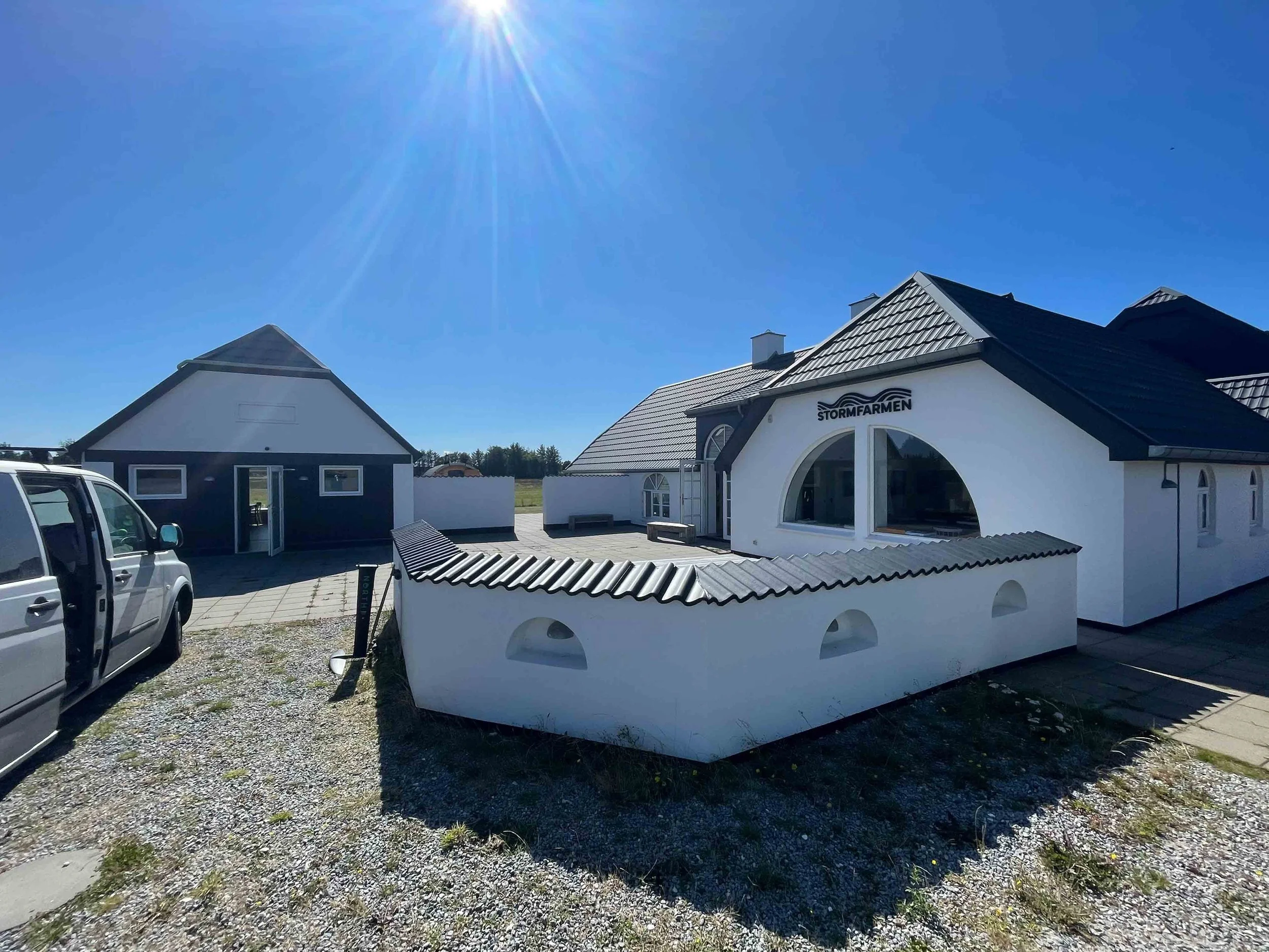 Modern weißes Gebäude mit dem Schriftzug 'STORMFARMEN' auf der Front, umgeben von einer kleinen weißen Mauer, eineparkende weiße Van, und eine offene Tür auf der linken Seite; sonniger Himmel mit Sonnenstrahlen.